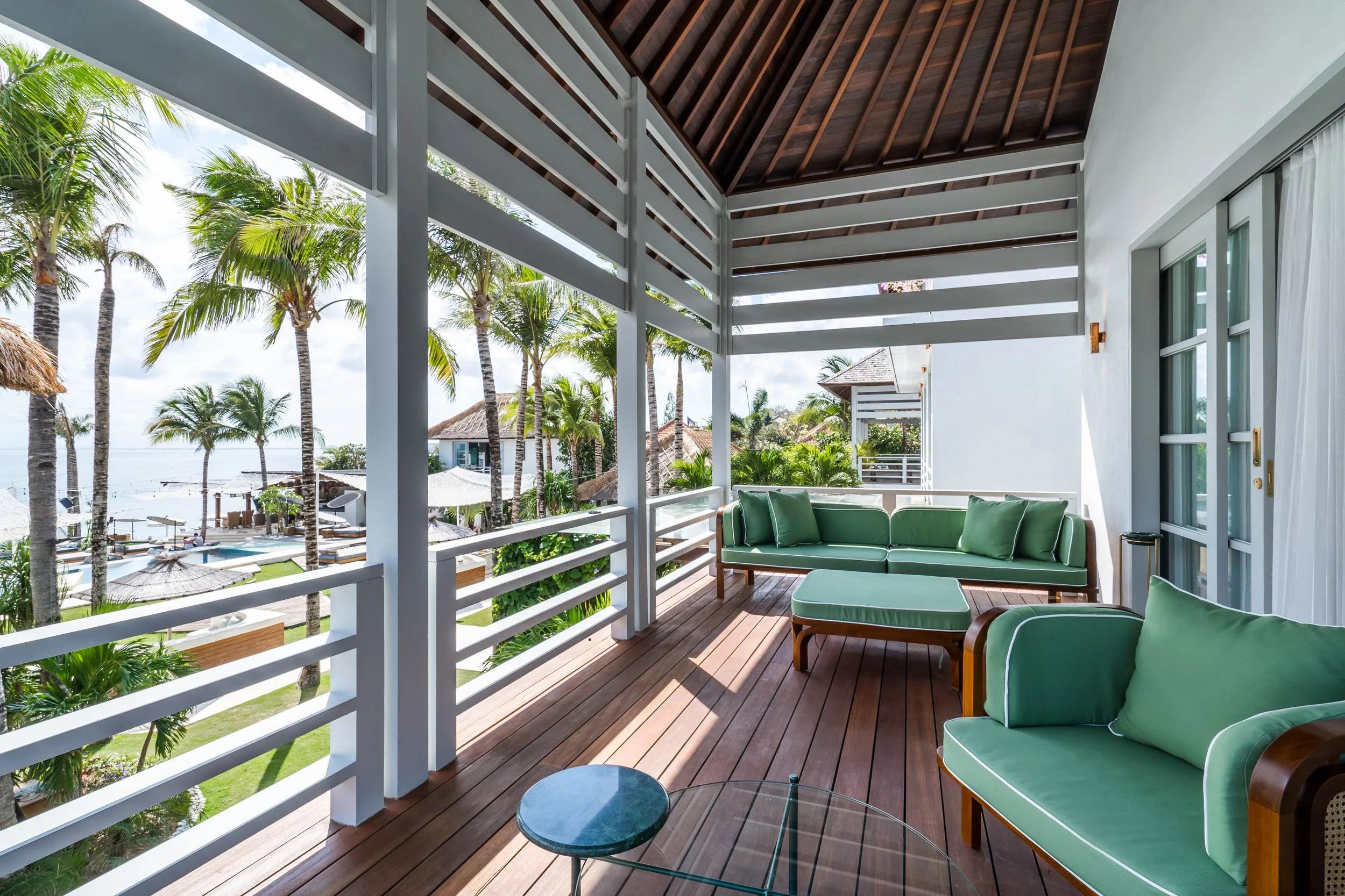 A balcony with green outdoor seating, a small glass table, white railings, and a view of palm trees, thatched umbrellas, and a pool area near the ocean.