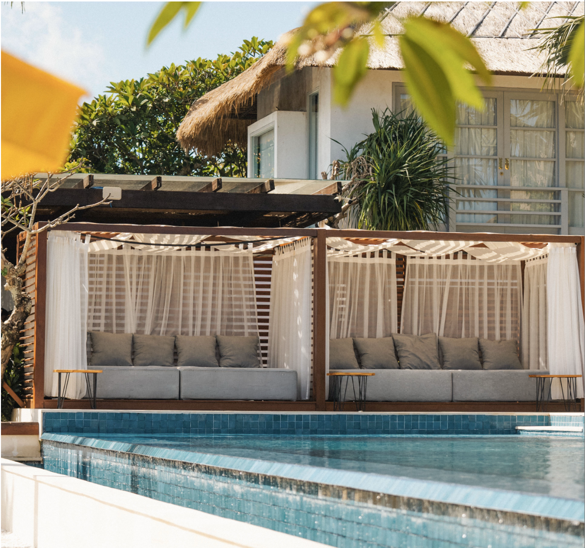 Luxury poolside cabanas with curtains and cushions, overlooking a swimming pool, with a tropical house and greenery in the background.