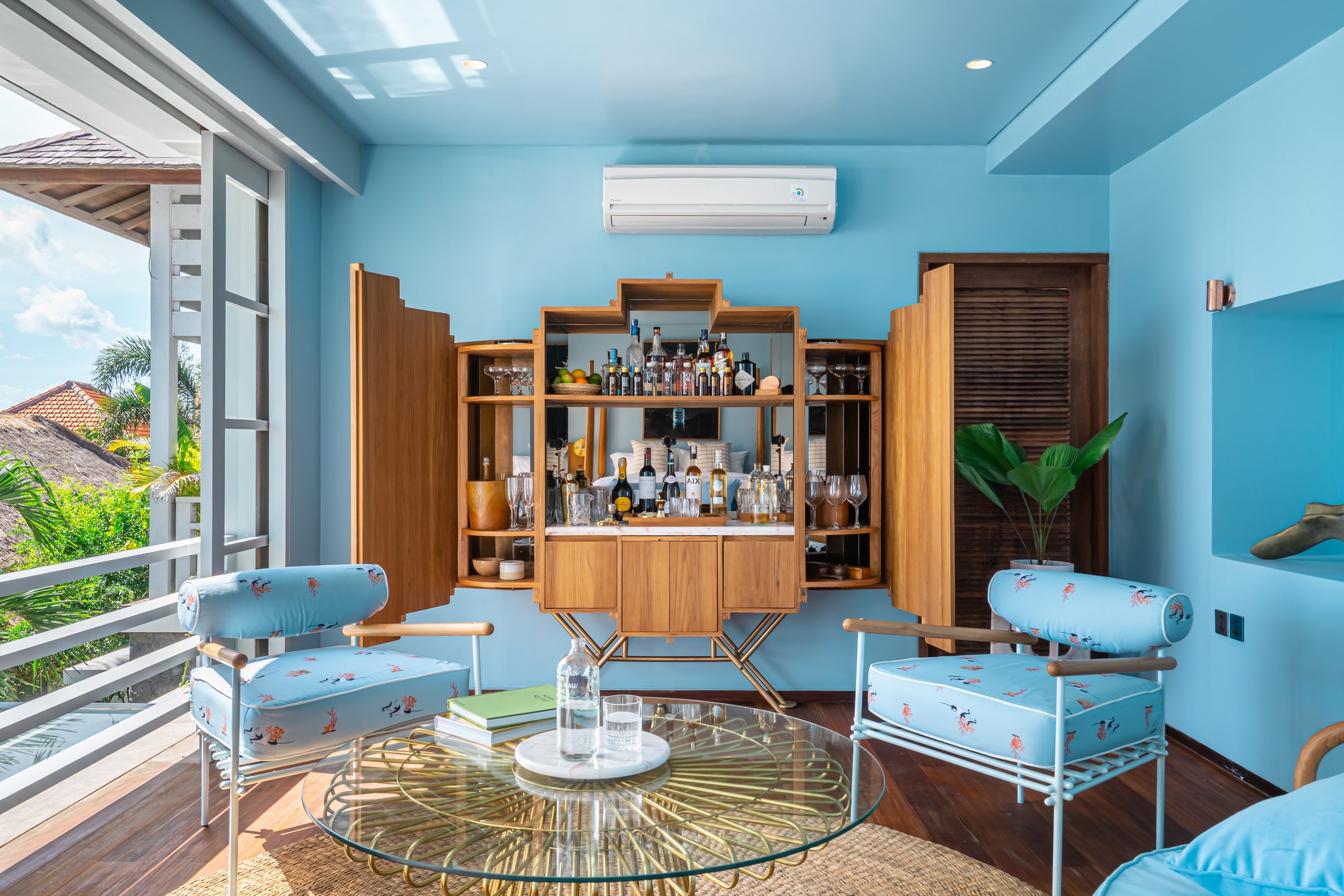 A balcony with two light blue armchairs with flamingo and tropical pattern, a glass-top coffee table with a water bottle and books, and a bar area with bottles and glasses, set against bright blue walls and a view of tropical trees and rooftops outsi