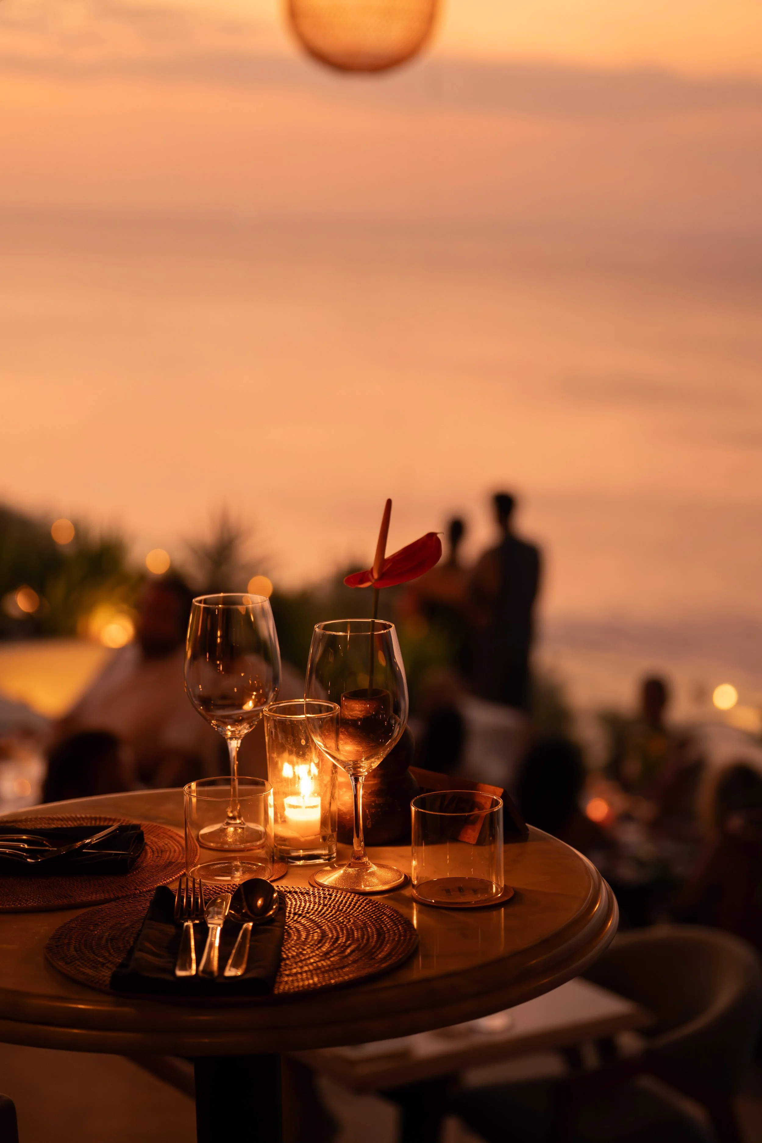 Elegant outdoor dinner table at sunset with wine glasses, candles, and a floral arrangement, overlooking a beach with people and a sunset sky in the background.