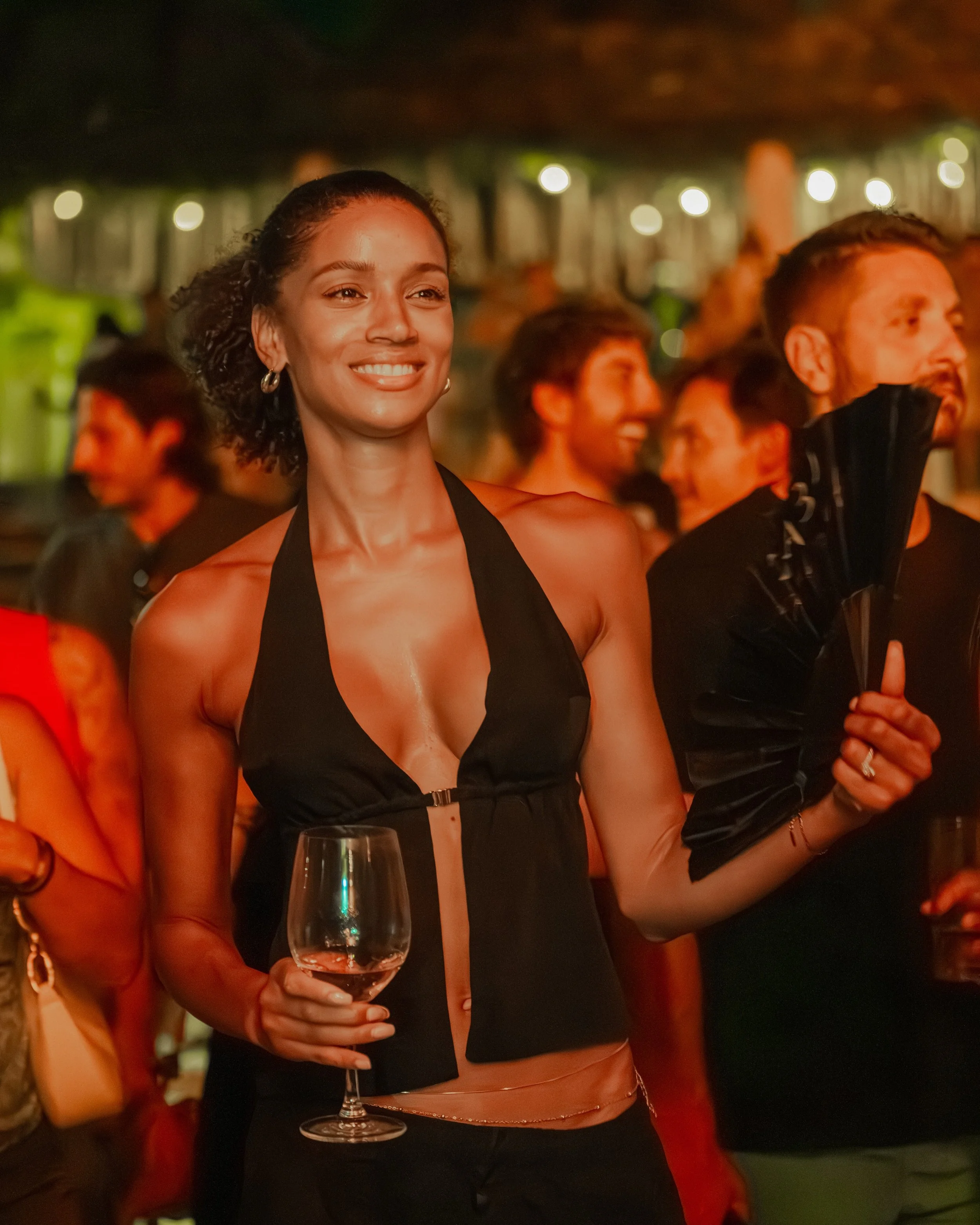 A woman in a black halter top holds a glass of rosé wine at a social event, smiling with other people in the background under warm lighting.