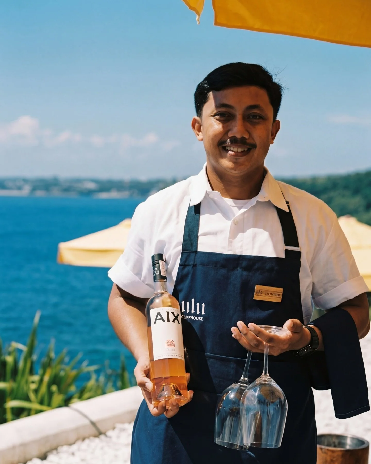 A smiling man dressed in a white shirt and dark apron holding a bottle of pink wine labeled 'AIX' and a wine glass, standing outdoors near a body of water with a yellow beach umbrella and green landscape in the background.