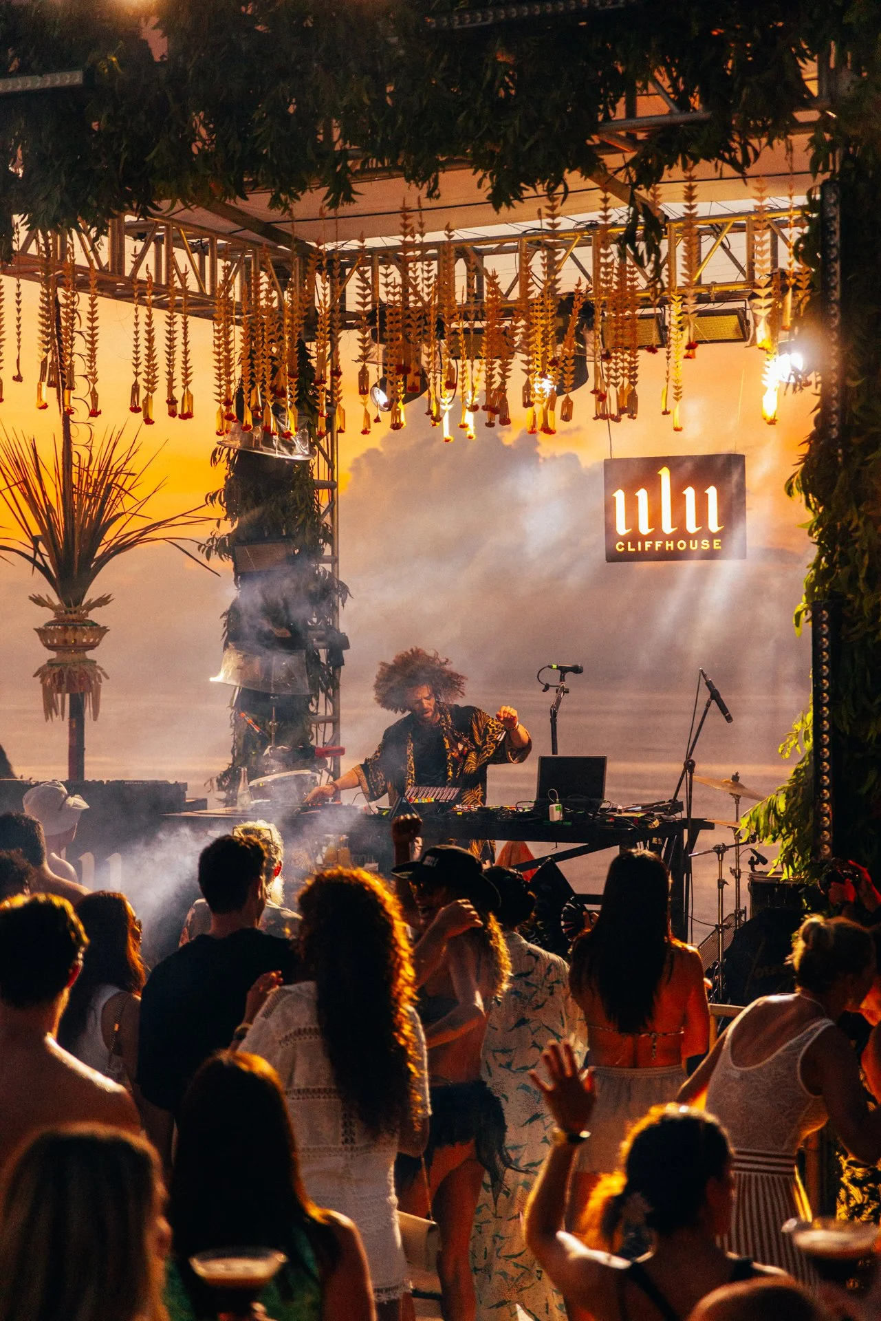 A DJ or electronic music performer with curly hair playing music on a stage at sunset, with an audience dancing in front of her at a beach party or outdoor event. The stage is decorated with hanging ornaments and palm trees, with the sign 'WLI Cliffhouse' illuminated in the background.