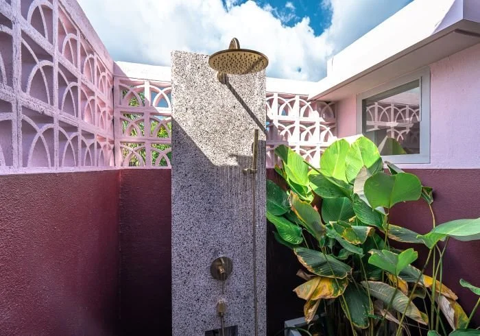 Outdoor shower with rain shower head on a granite wall, next to large green tropical plants and pink wall with decorative concrete blocks.