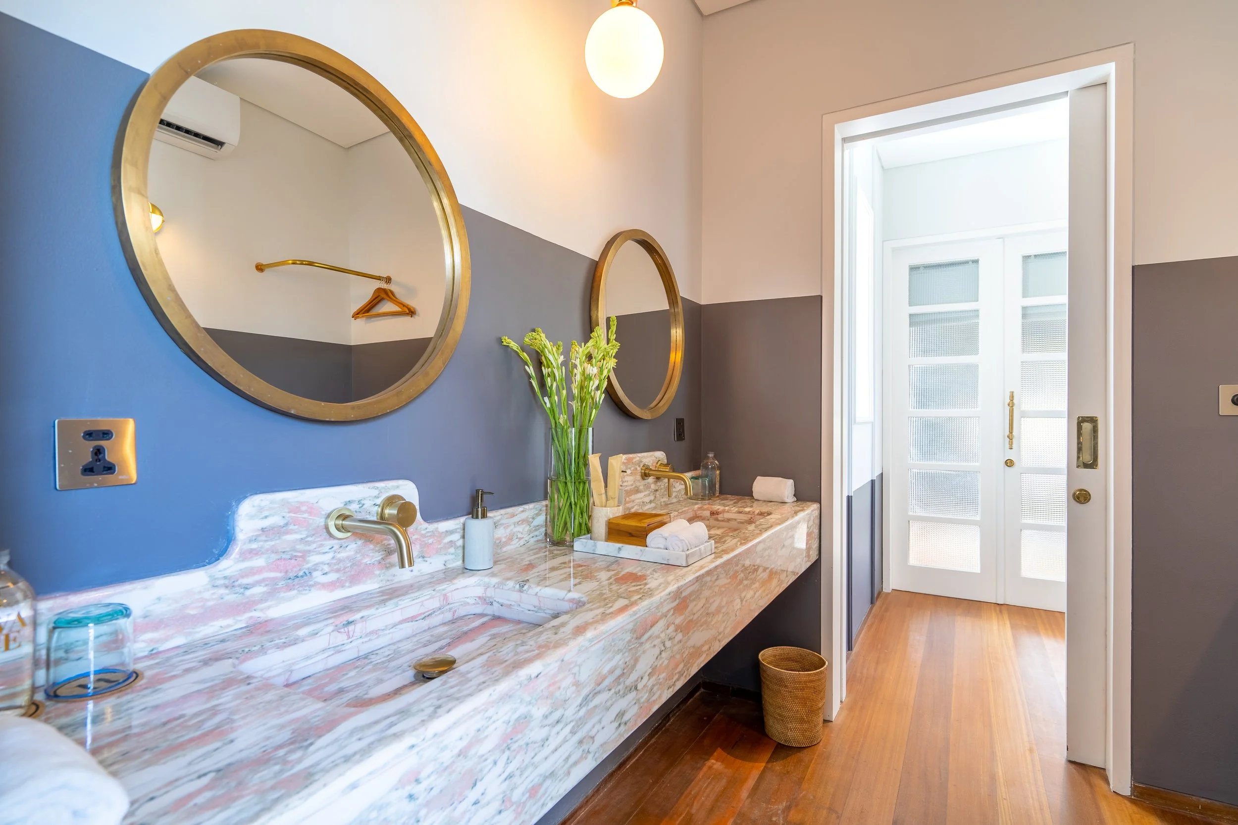 Modern bathroom with blue and gray walls, featuring two round mirrors, a marble countertop with a sink, a vase with green flowers, and a door leading outside. The bathroom has wooden flooring and is decorated simply and elegantly.