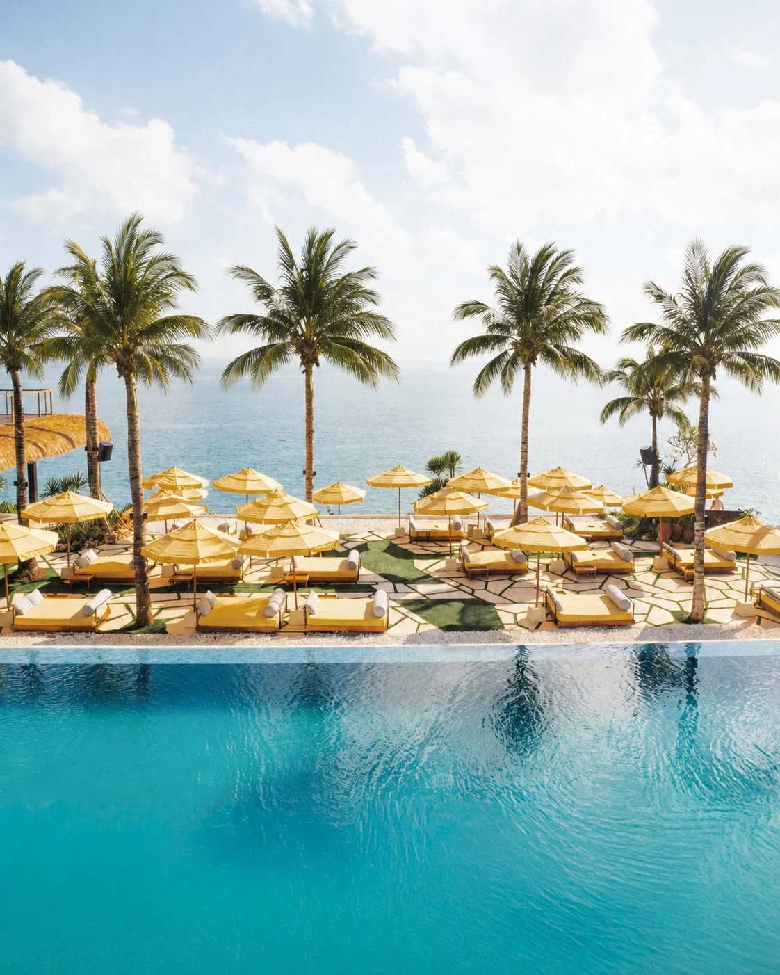 View of a luxurious pool area with yellow lounge beds and umbrellas, overlooking the ocean with tall palm trees and a cloudy sky.