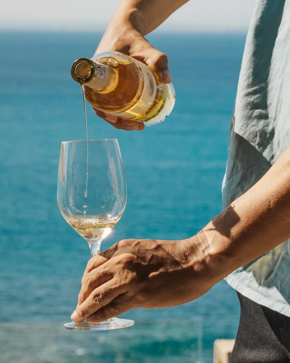 Person pouring white wine into a wine glass with a blue ocean background.