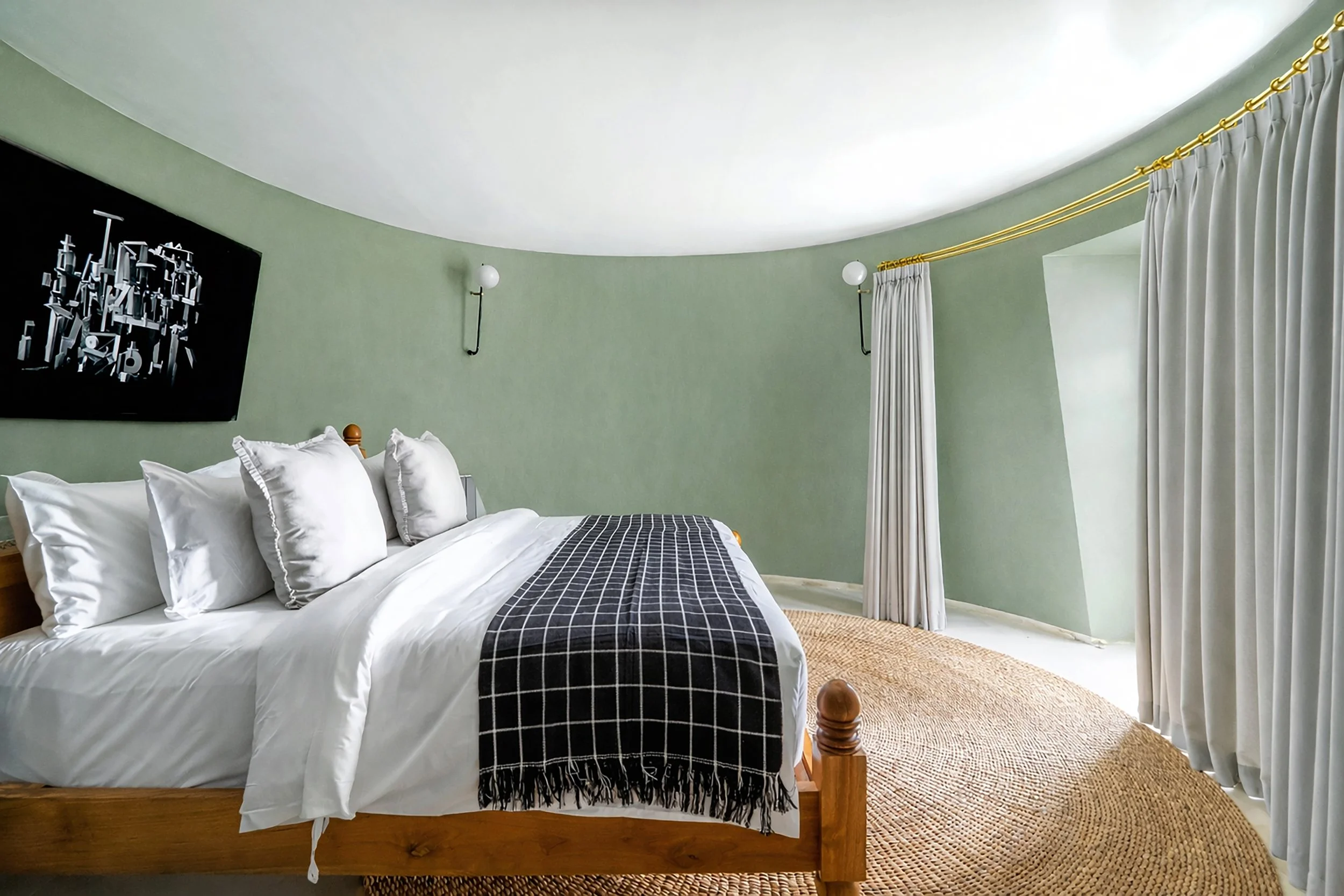 A bedroom with a wooden bed frame, white bedding, several white pillows, and a black and white checkered blanket. The room has light green walls, two wall-mounted lamps, and a window with white curtains and gold curtain rods. There is a black abstrac