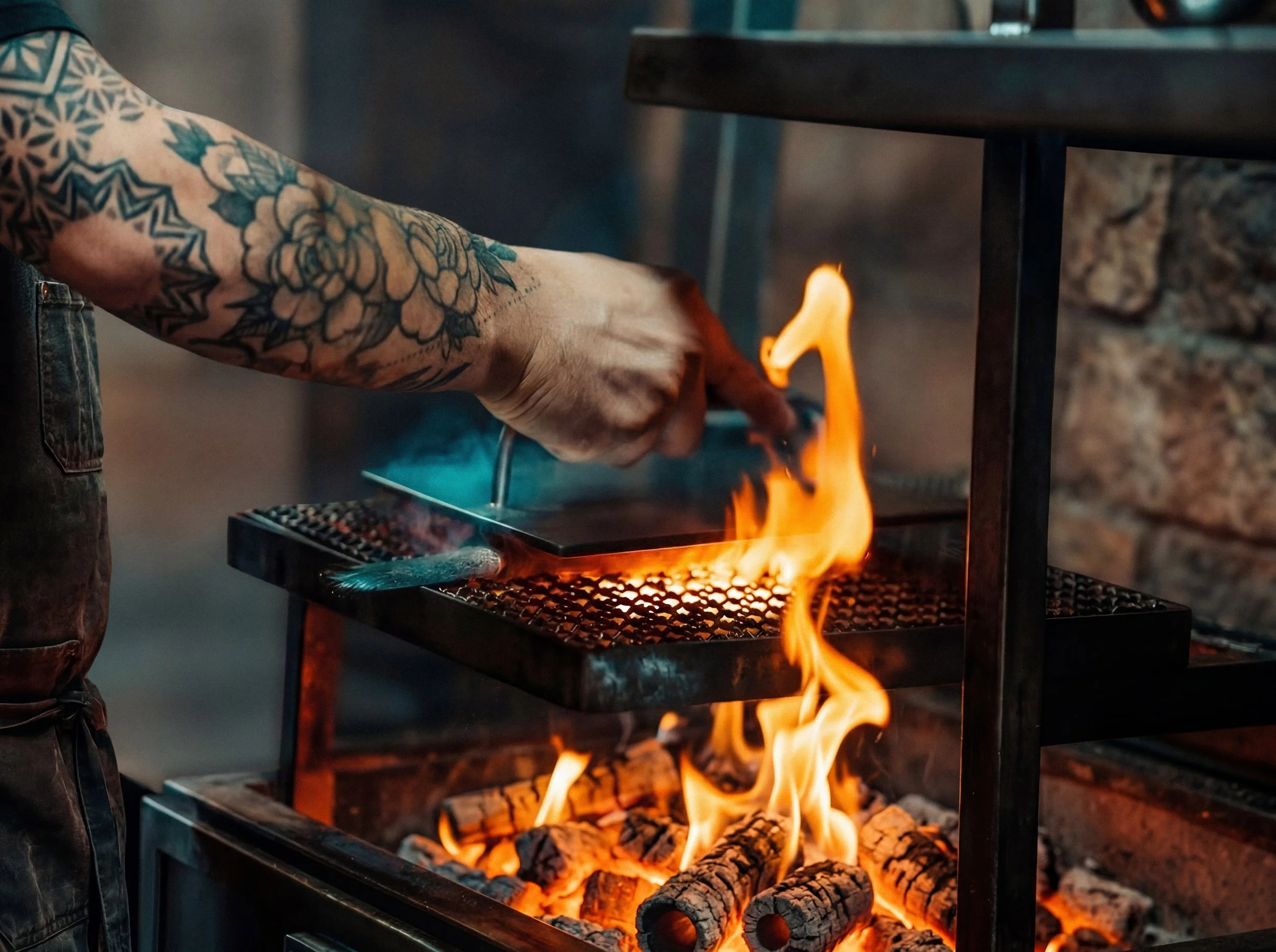 A person with a tattooed arm reaching towards a grill with flames, cooking over wood and charcoal.
