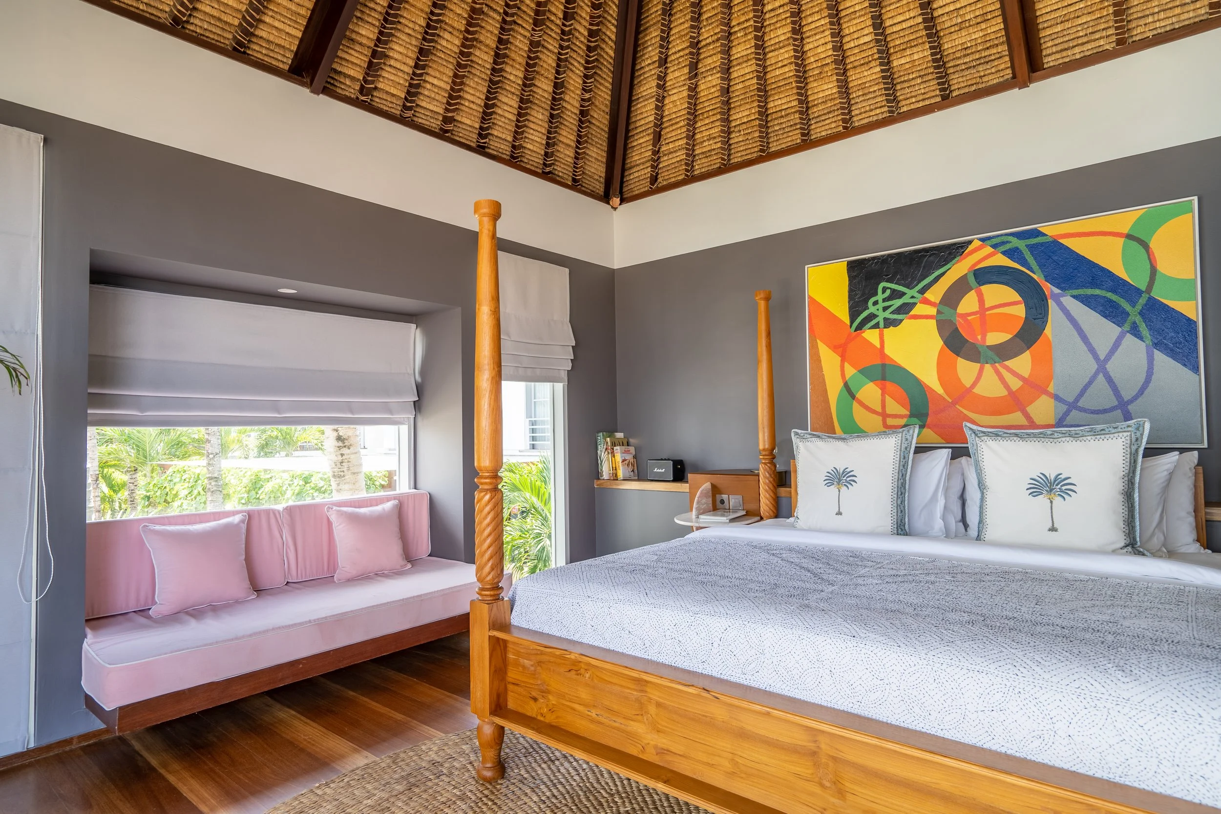 A bedroom with a wooden four-poster bed, white bedding, and tropical-themed pillows. A pink cushioned window seat with pillows is beneath a window with white blinds, and a colorful abstract painting hangs on the gray wall. The ceiling is thatched.