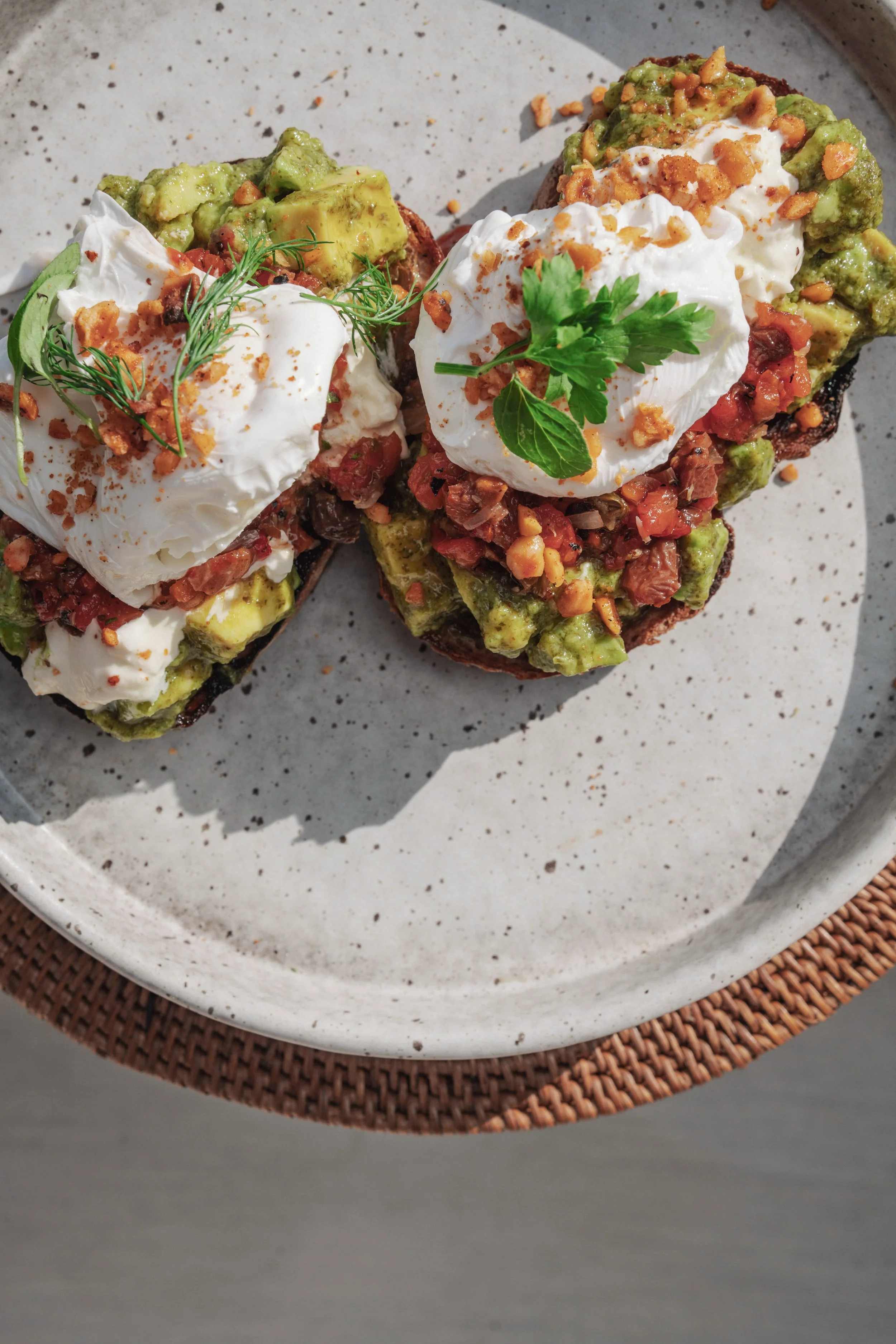Two open-faced breakfast tostadas topped with sour cream, chopped tomatoes, guacamole, and garnished with fresh herbs, serving on a speckled white plate placed on a woven placemat.
