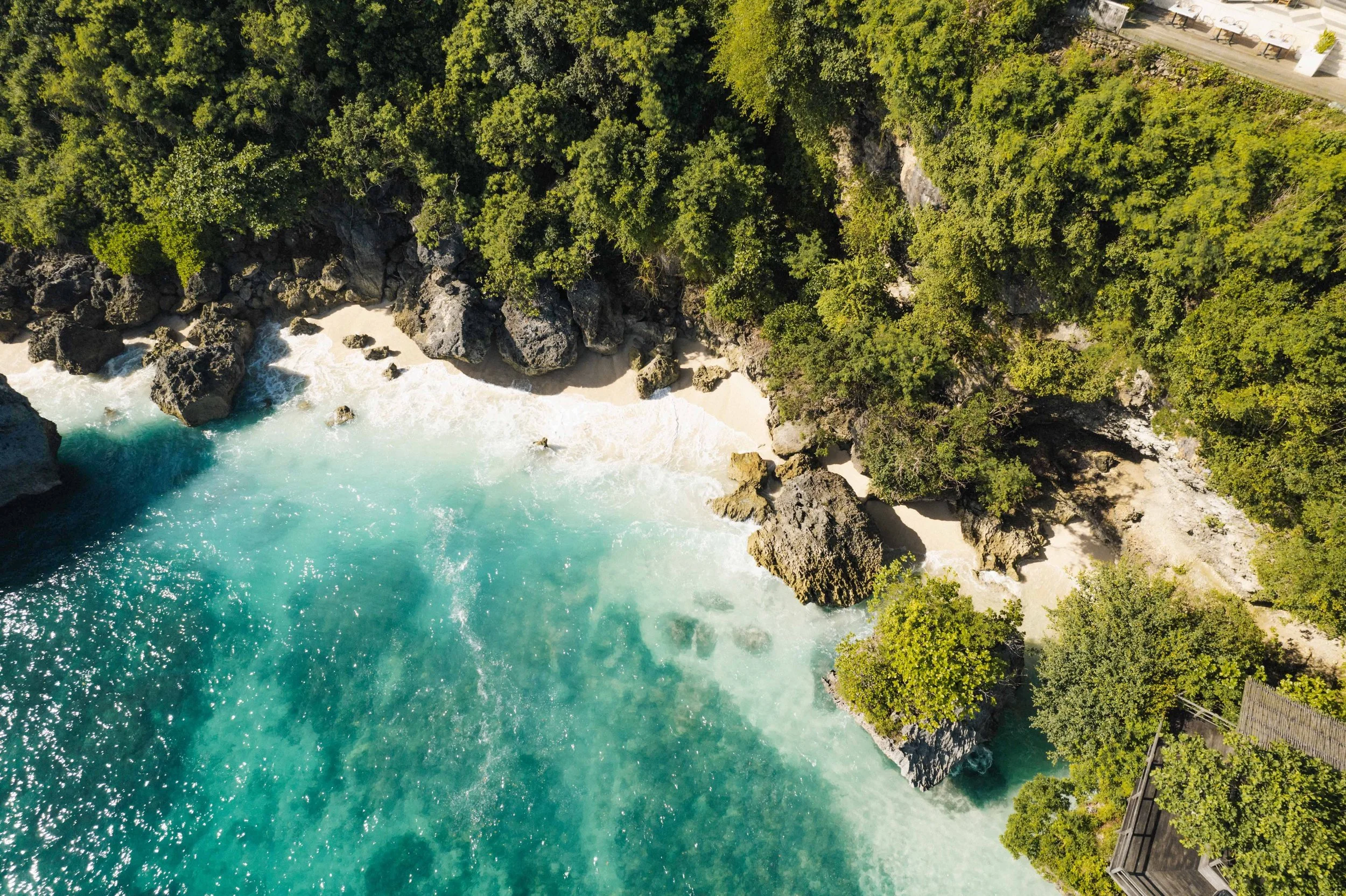 Aerial view of the ocean with turquoise water crashing against rocky cliffs covered in lush green trees, and some outdoor seating area visible at the top right corner.