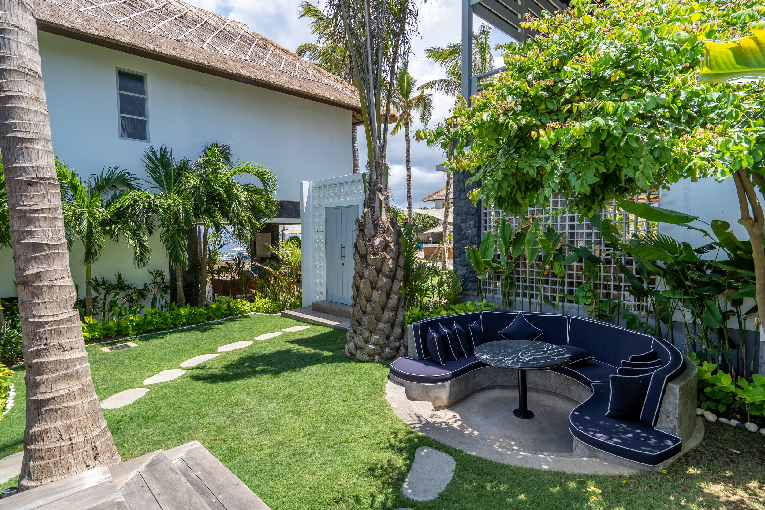 Tropical outdoor lounge area with black curved sofa and marble table, surrounded by palm trees, lush green bushes, and a white building with thatched roof in the background.