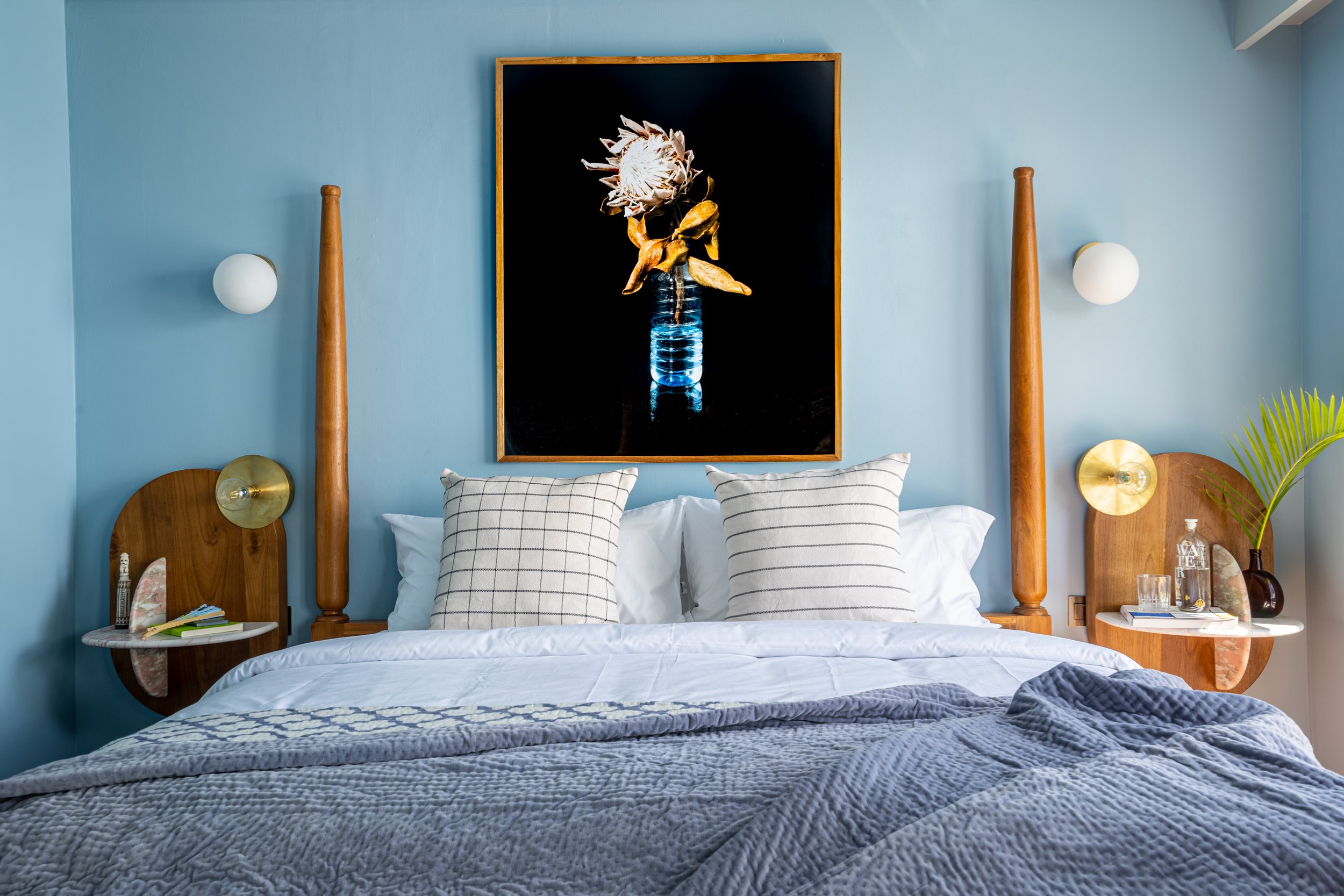 A bedroom with blue walls, featuring a large framed artwork of a flower in a glass vase hanging above the bed. The bed has white pillows with black stripes, and a throw blanket at the foot. Two wooden headboards with attached round bedside tables sup