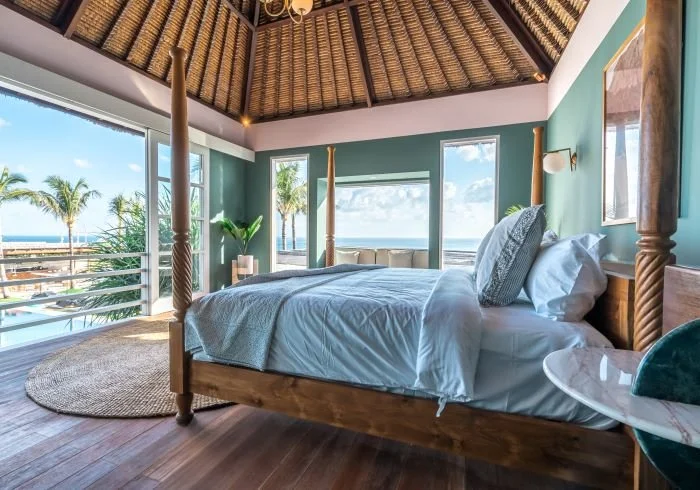 A tropical bedroom with a four-poster bed, large windows with ocean views, palm trees outside, and a high thatched ceiling.