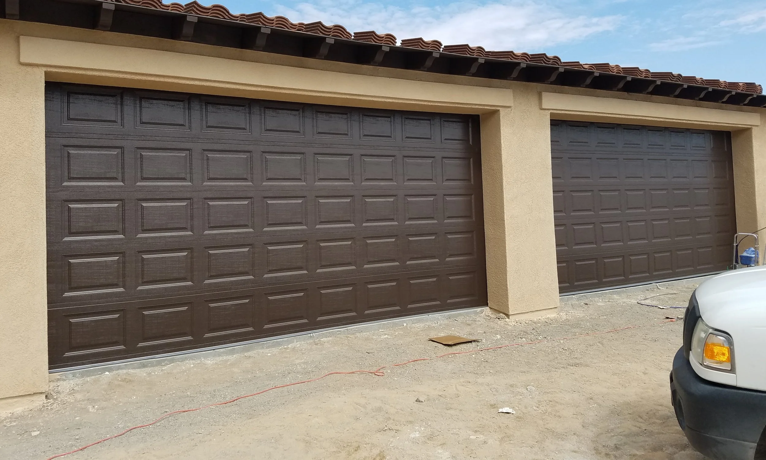 A row of two closed brown garage doors on a beige stucco house, with a car partially visible in the foreground on the right side and a blue sky with some clouds above.