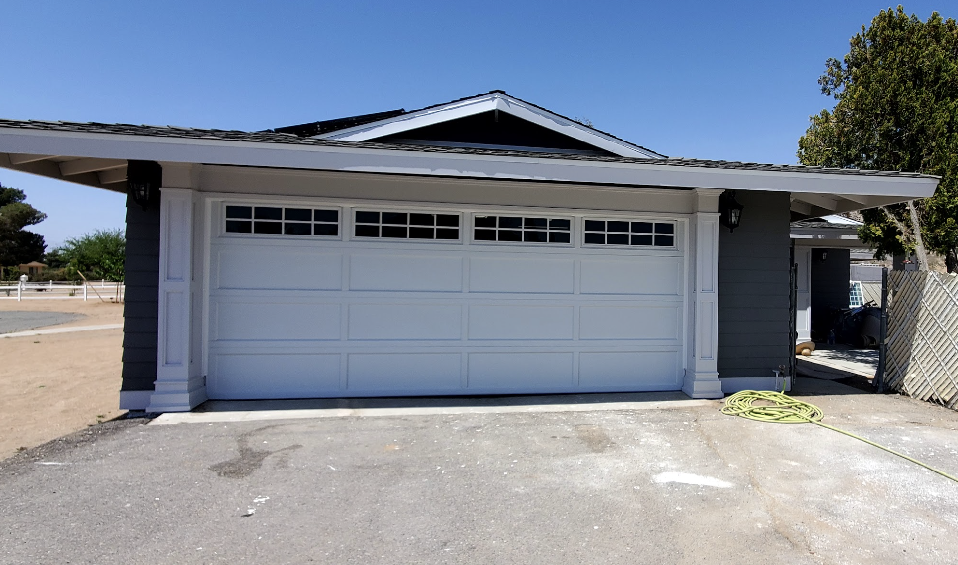 A residential garage with a white door and a small row of windows at the top. The house has dark gray siding with white trim. A black lantern-style light is mounted on the corner of the house near the garage, and a green garden hose is coiled on the 