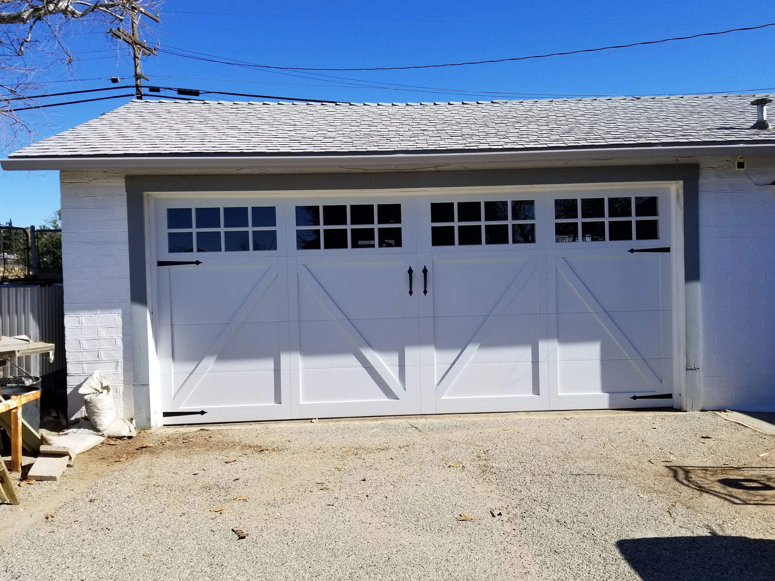 A white garage door with black hardware, set in a white brick wall, with a gravel driveway in front. The roof is shingled, and there are utility wires overhead against a clear blue sky. There are some construction materials and debris on the left sid