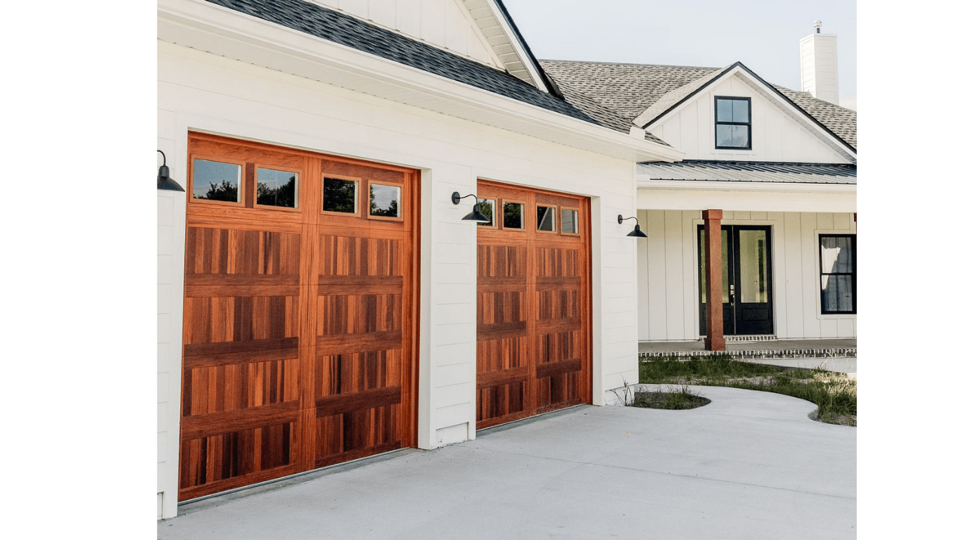 Two wooden garage doors with small windows at the top, attached to a white house with a front porch and black front door.