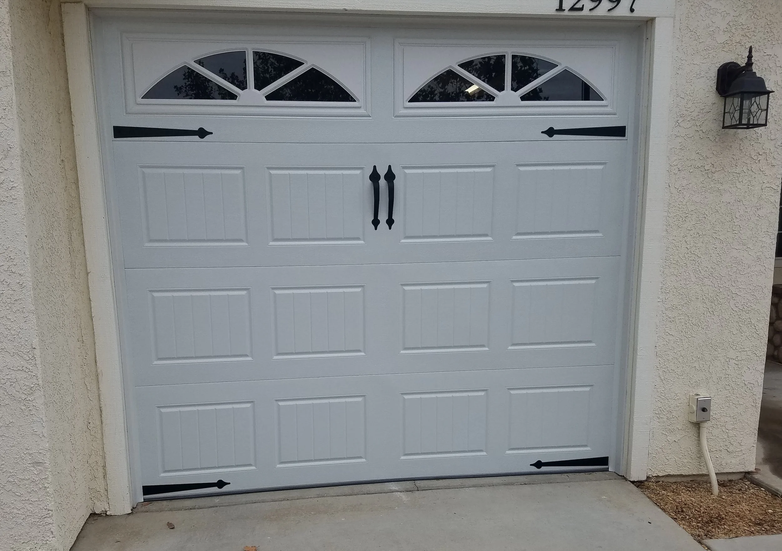 White garage door with decorative black hinges and handles, located on a beige stucco house. A black outdoor wall light fixture is mounted to the right of the garage, and there is a small utility box and an electrical conduit on the right side.