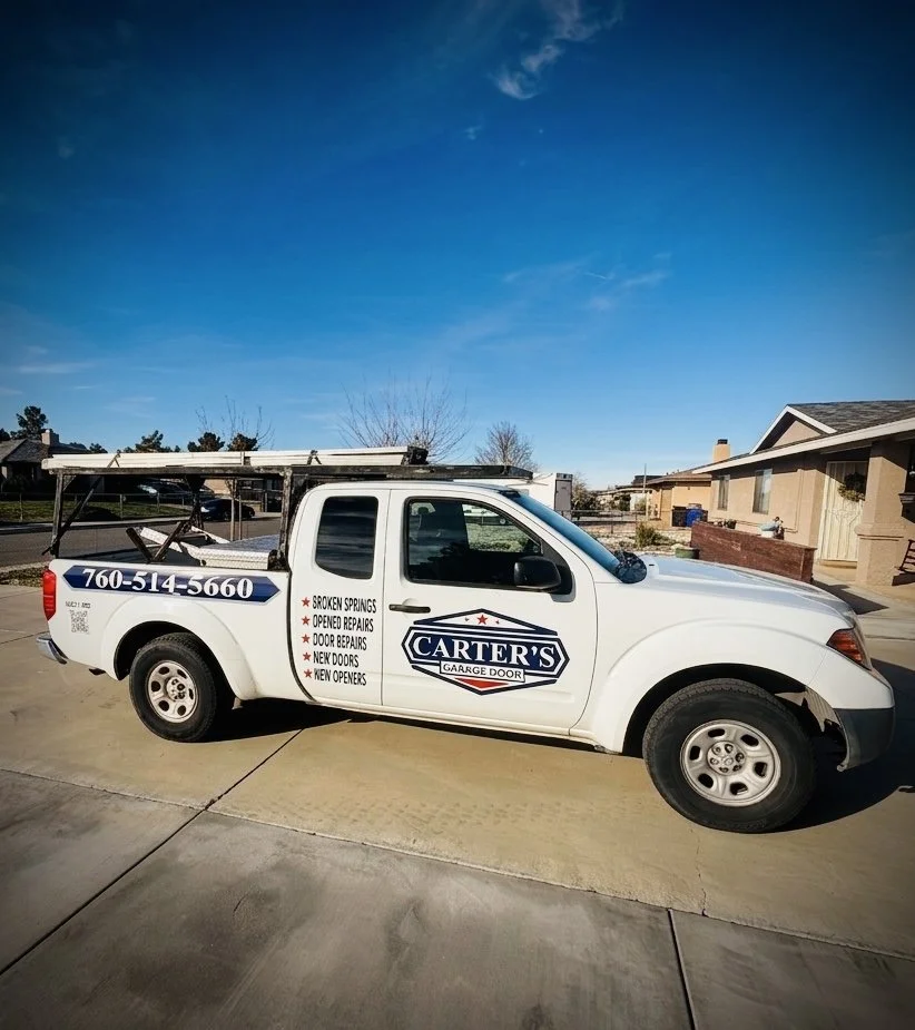 A white service truck parked on a driveway in front of suburban houses under a clear blue sky. The truck has the logo for Carter's Garage Door and lists services such as broken springs, opener repairs, door repairs, new doors, and new openers.
