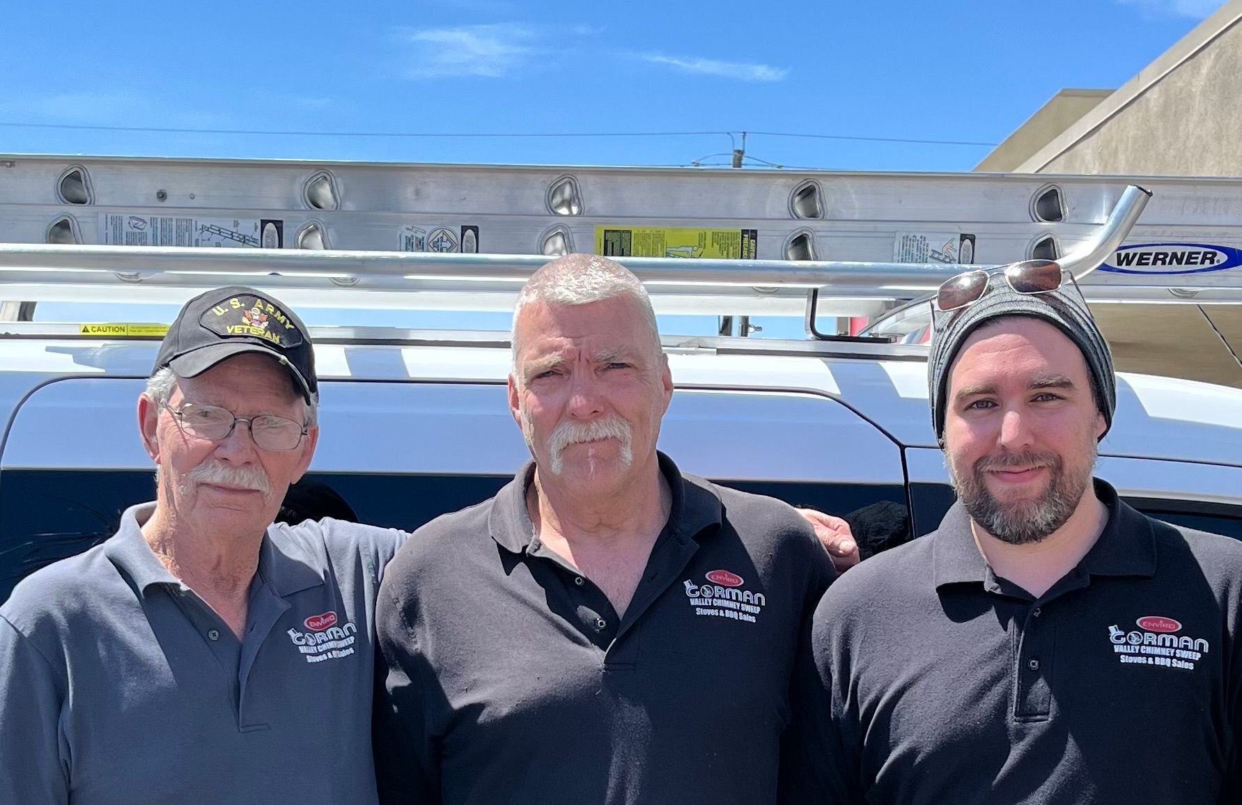 Three men standing side by side outdoors with a vehicle and ladder in the background. All are wearing black or gray polos with a logo that says 'Cormac Valley Chimney Sweep Stoves & BBQ Sales.'