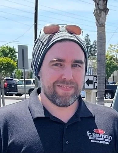 Man with a beard and wearing a striped headscarf with sunglasses on top, standing outdoors near parked cars and trees.