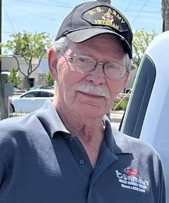 An elderly man wearing glasses, a U.S. Army Veteran cap, and a navy blue polo shirt with a logo, standing outdoors near a vehicle.