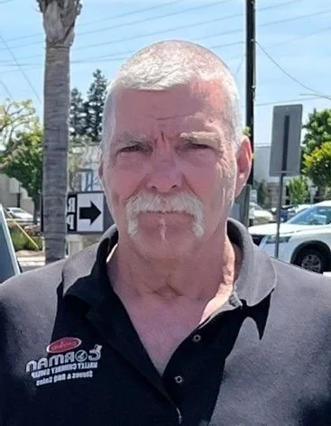 A middle-aged man with short gray hair and a mustache wearing a black collared shirt with a logo, standing outdoors on a sunny day, near trees, cars, and electrical poles.