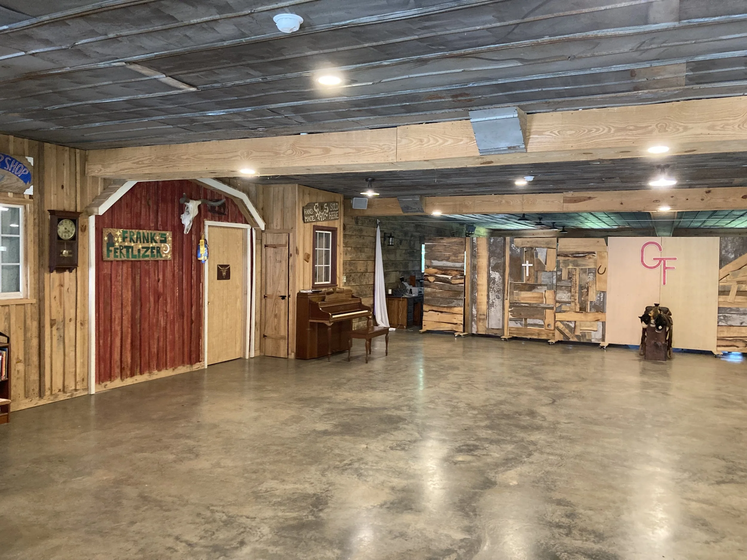The interior of a rustic lodge or barn, with wood-paneled walls and ceiling, some lighting fixtures, and a concrete floor. There are various signs and decorative items on the walls, including a sign reading 'Frank's Fertilizer' and a cow skull mounted above a door.
