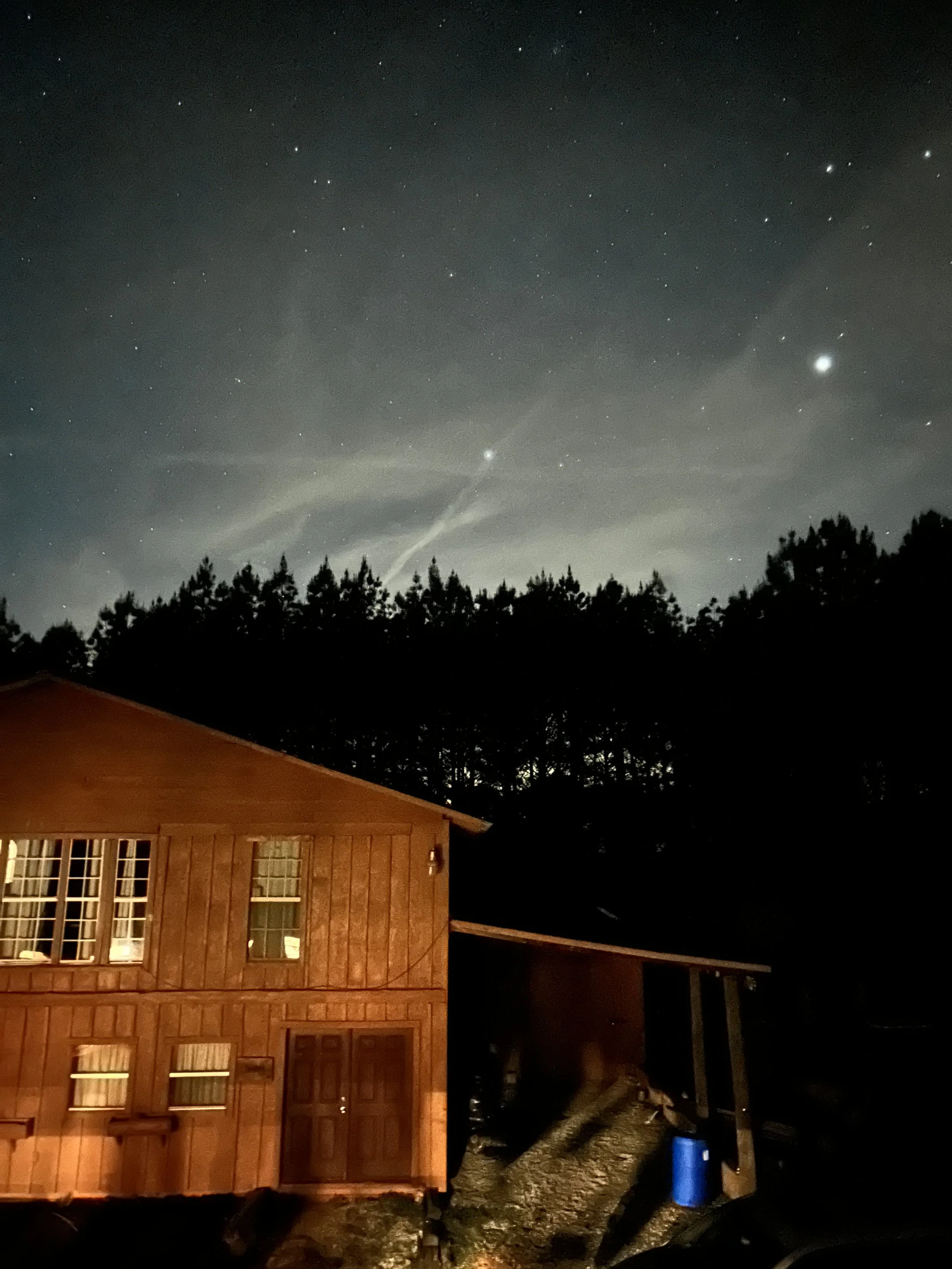 Nighttime view of a house with illuminated windows and a wooden exterior, a large tree in the yard, and a sky filled with stars, including a bright planet and a streak of light