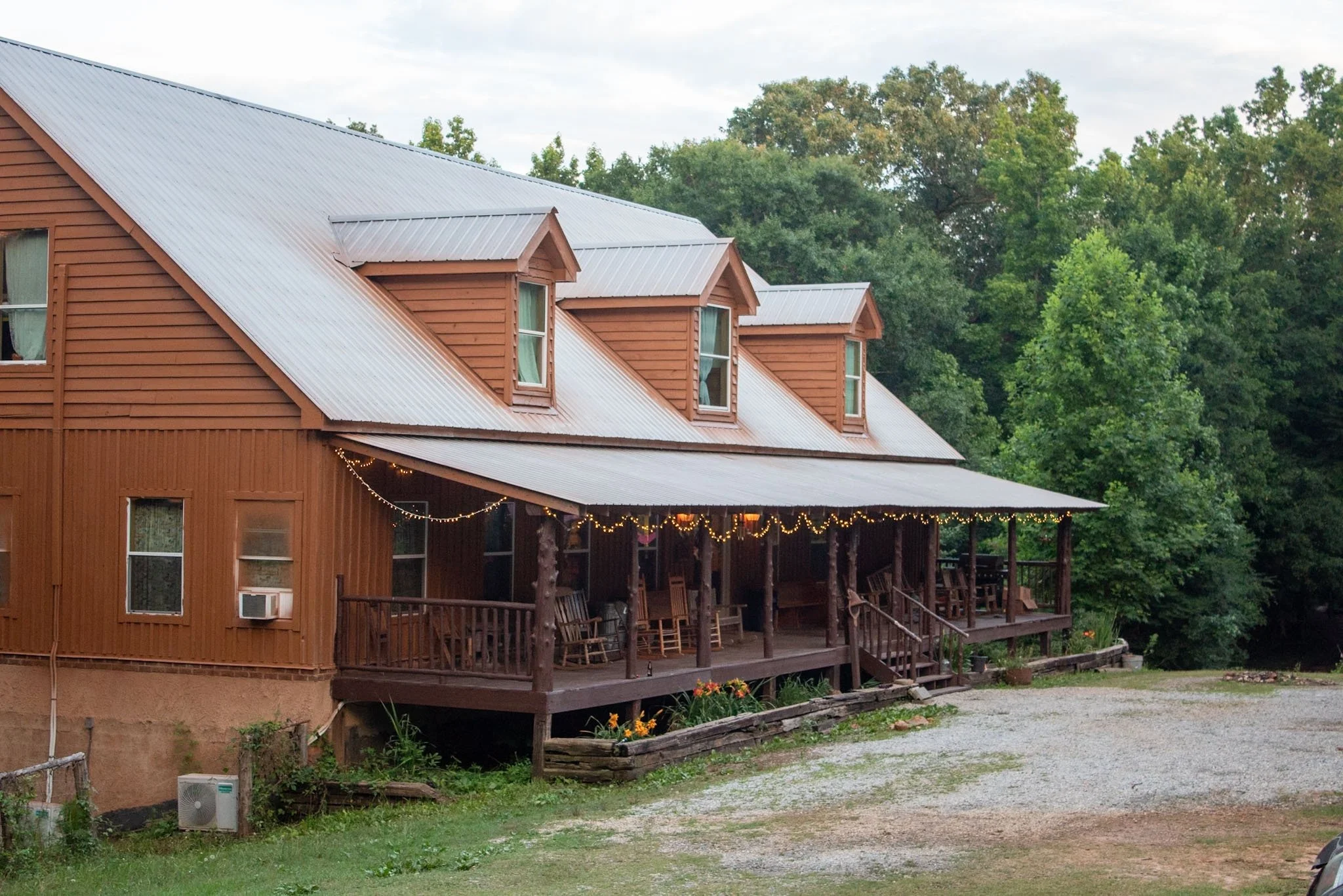 A two-story wooden building with a metal roof and multiple dormer windows, surrounded by green trees. The building has a balcony with string lights and chairs, and a gravel pathway in the foreground.