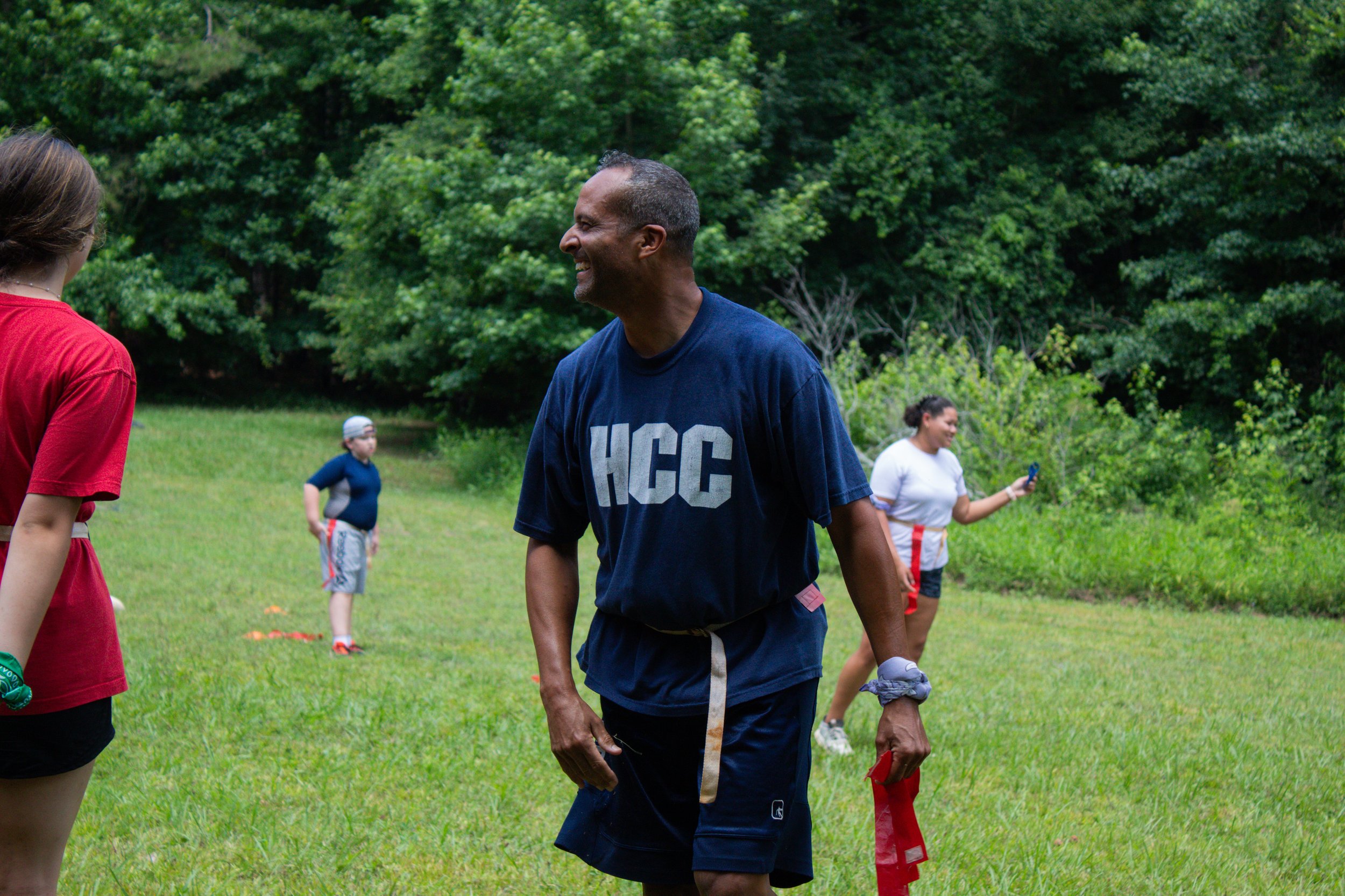 A man in a navy blue shirt with "HCC" printed on it, smiling and talking to a woman in a red shirt, in an outdoor park with lush green trees, children playing in the background.