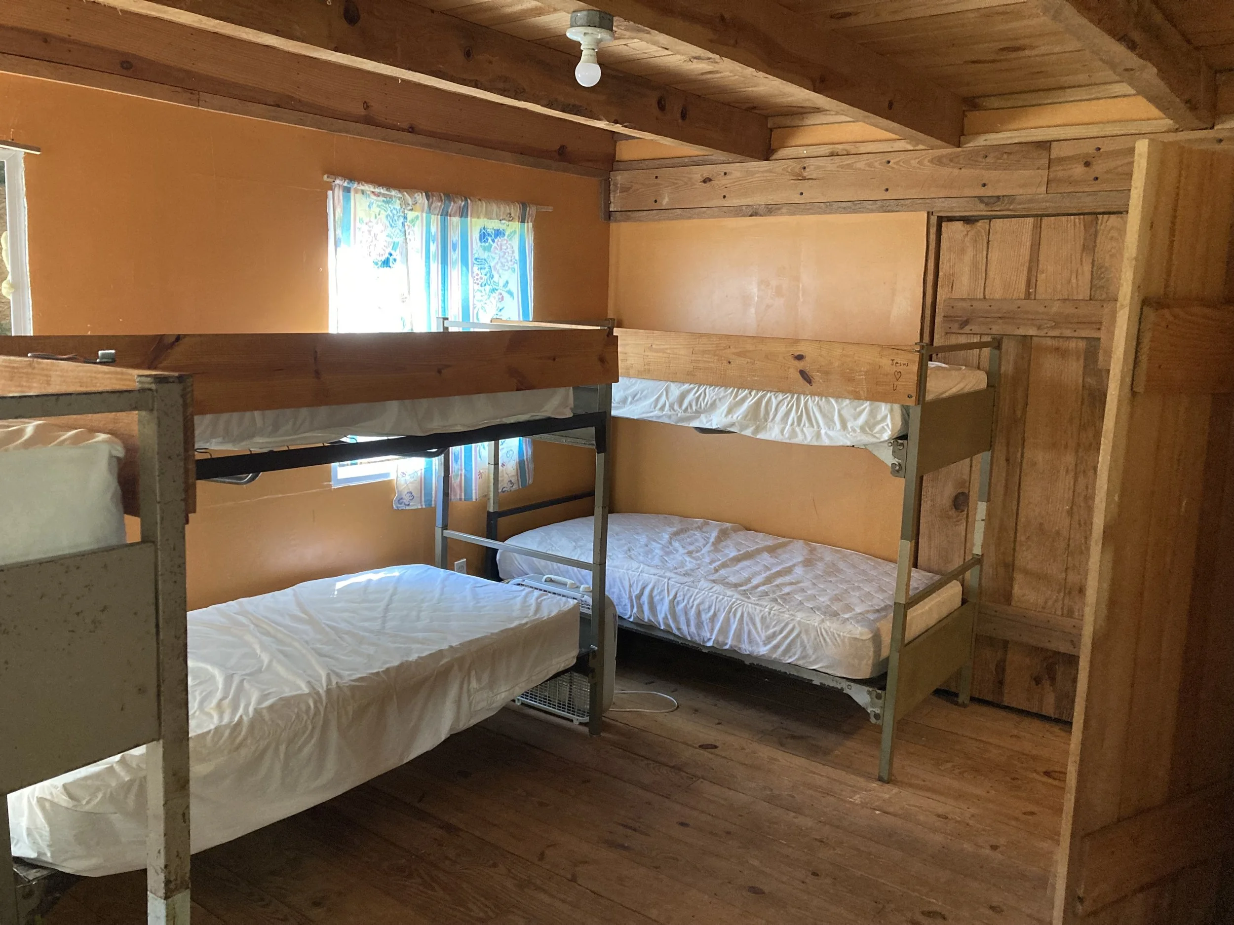 A rustic room with wooden walls and floors, featuring three silver bunk beds with white mattresses and a window with colorful curtains.