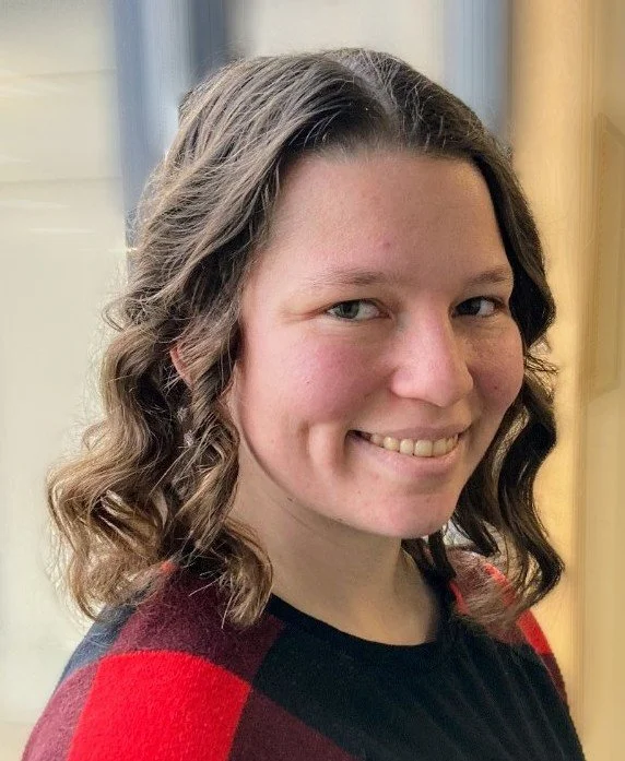 A woman with curly brown hair smiling, wearing a red and black top, standing indoors near a window.