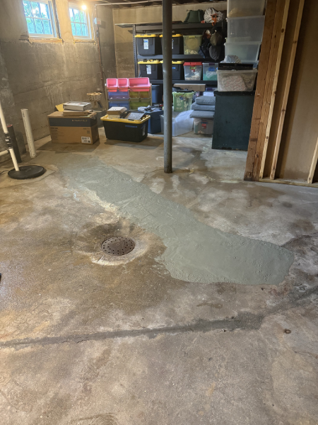 A basement or garage with a concrete floor that has a fresh patch of cement or concrete repair around a drain. There are various storage boxes and shelves along the wall, and a support pole in the center.