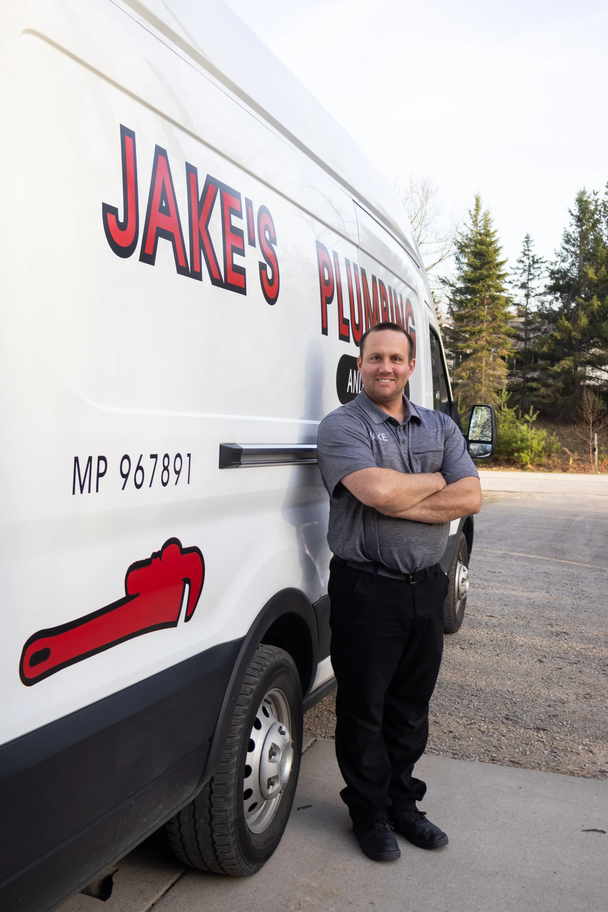 A man in a gray polo shirt and black pants standing with arms crossed in front of a white plumber's truck. The truck has the words 'Jake's Plumbing' in large red and black letters, and some images of a wrench and a pipe. Tall pine trees are in the background during daylight.