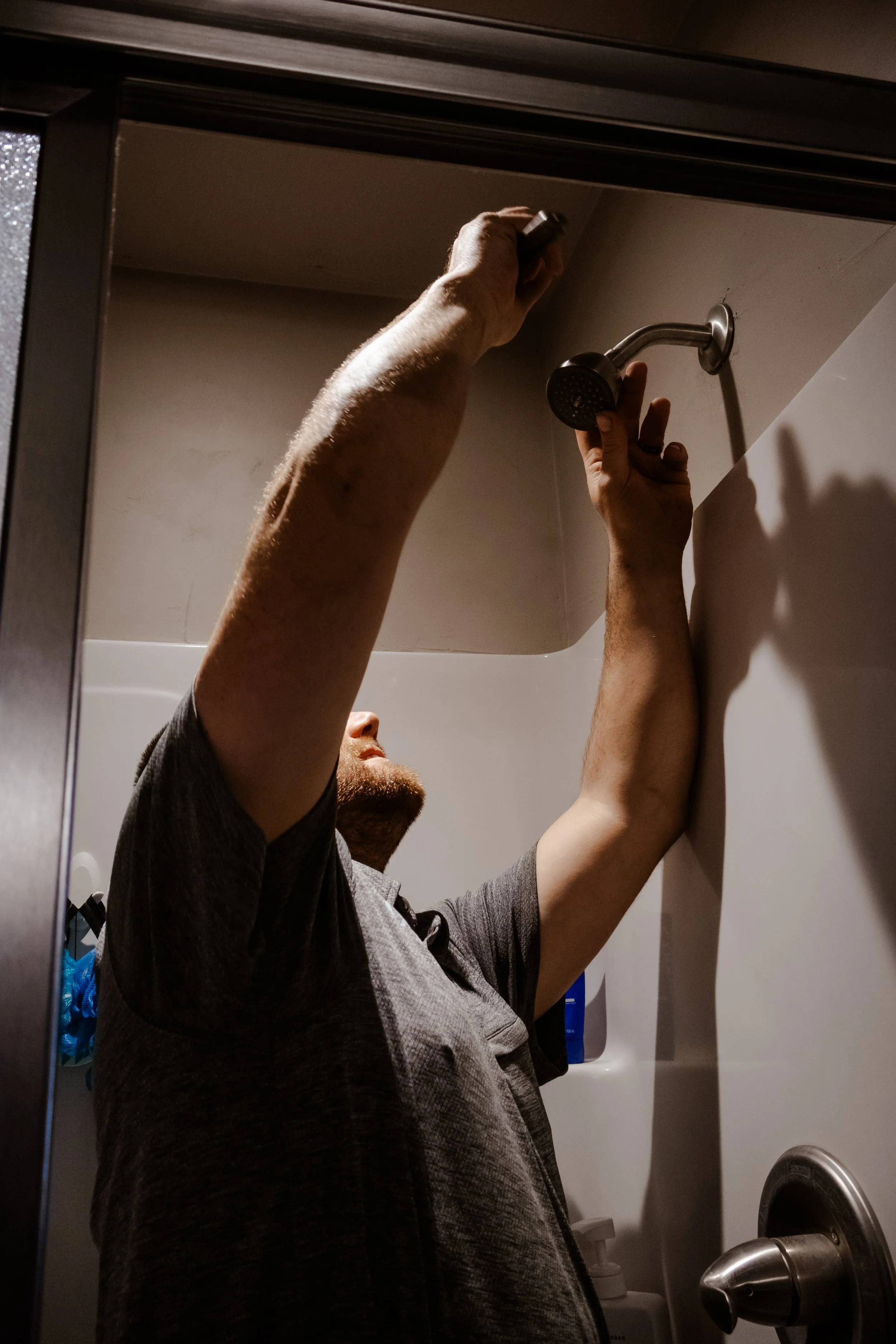 A person taking a shower in a wall-mounted shower with beige tiles, with their arms raised and hands gripping the showerhead, visible through a bathroom mirror.