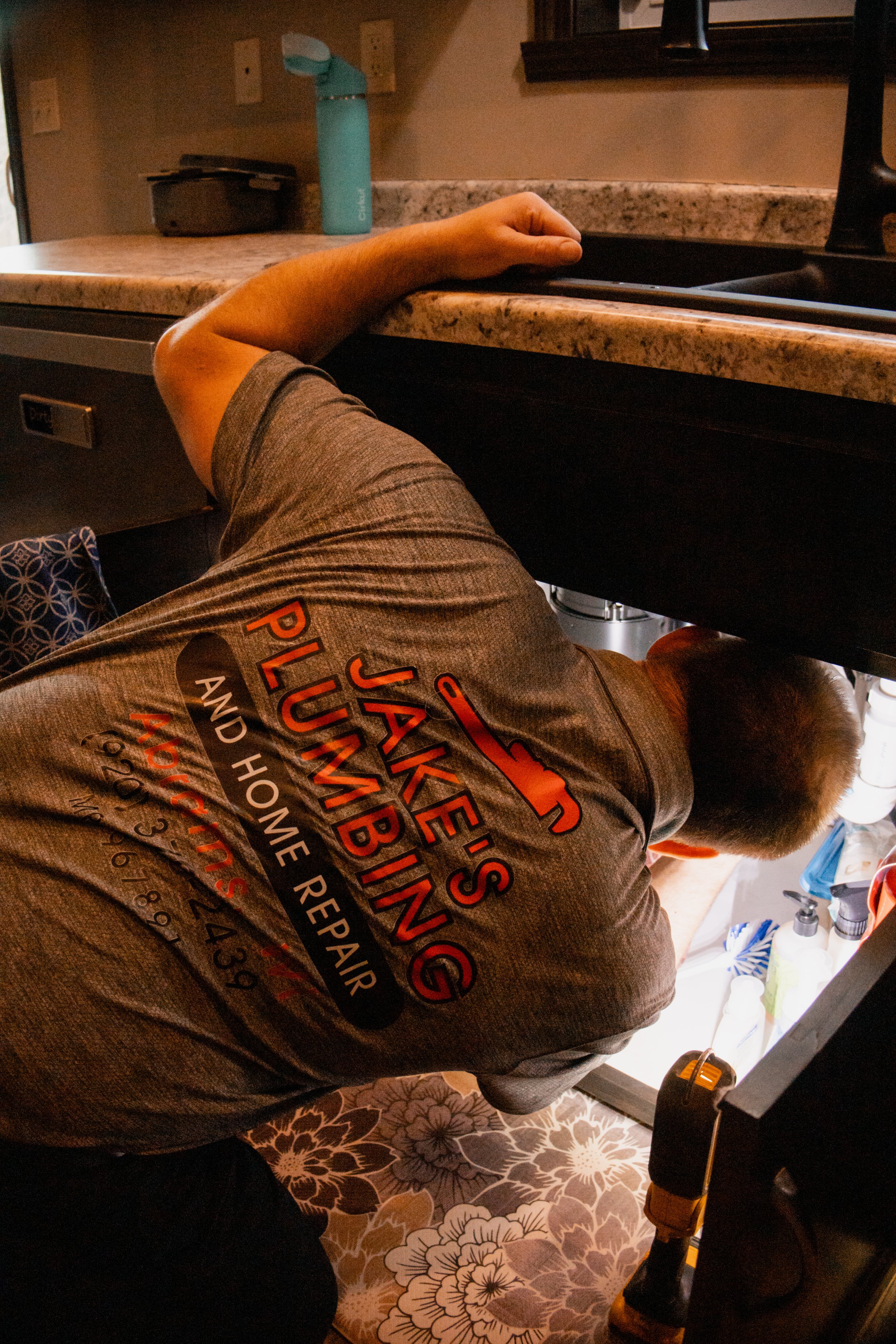 A person bend over at a kitchen sink, wearing a gray T-shirt with orange text, with various items on the counter and floral patterned flooring visible.