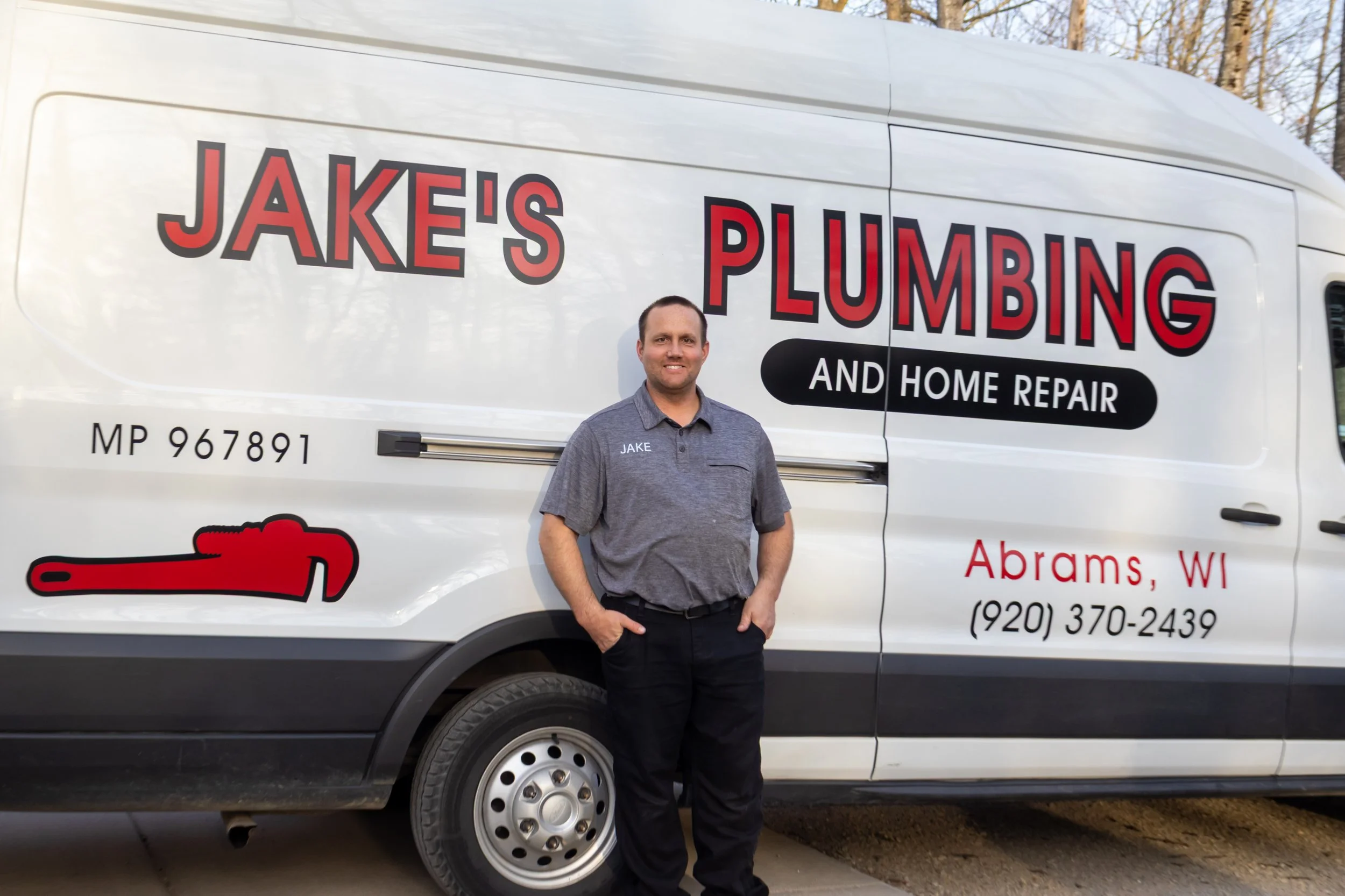A man standing in front of a white service van with red and black lettering. The van displays the company name 'Jake's Plumbing and Home Repair' and includes a red pipe wrench graphic. The man is wearing a gray shirt with the name 'Jake' embroidered on it and black pants, smiling at the camera.