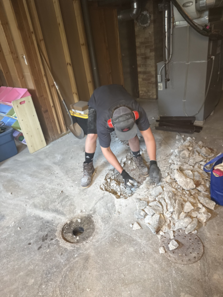 Person wearing gloves, a cap, and a mask, breaking concrete with a tool in a basement or garage.