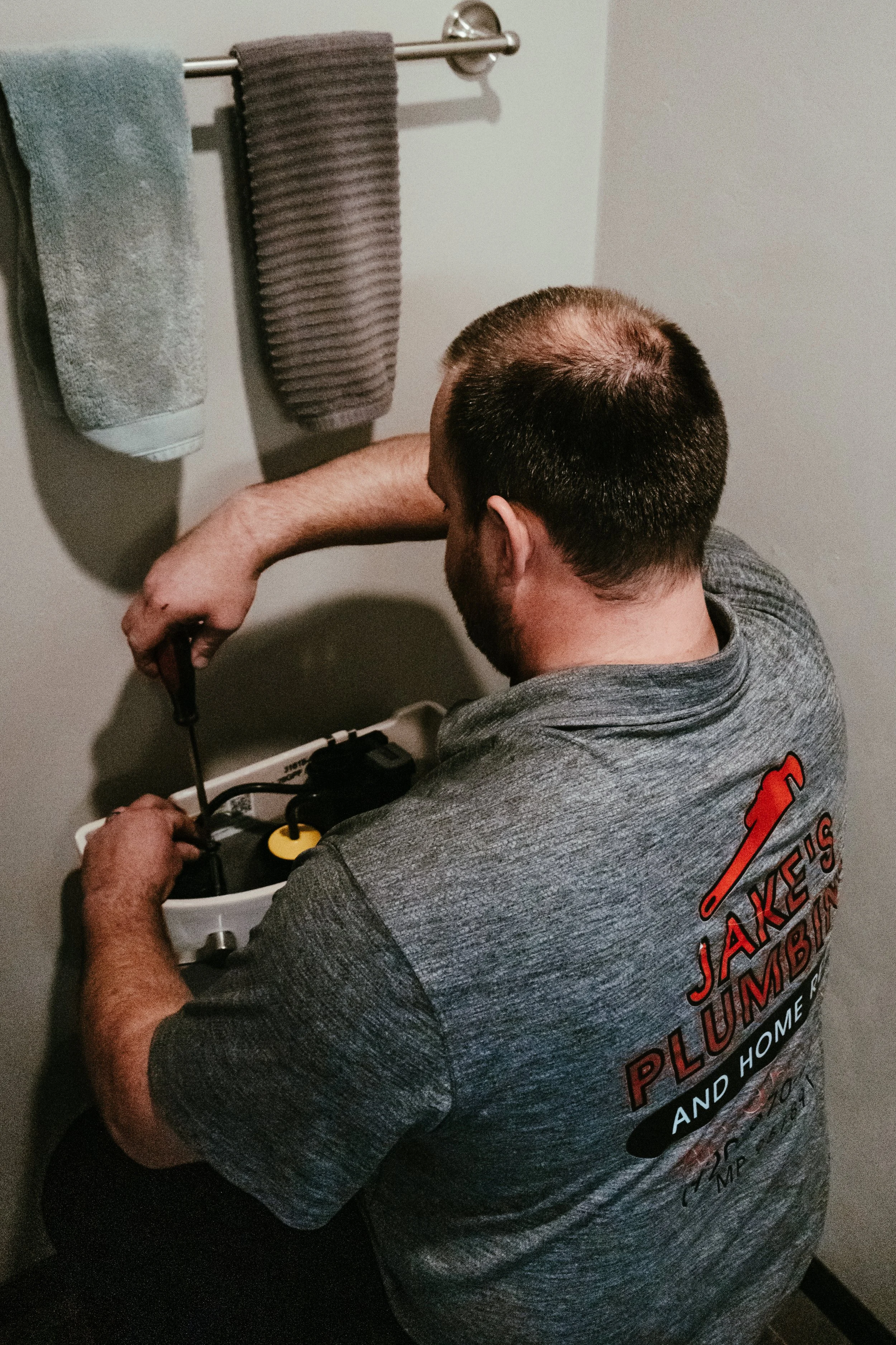 A man with short dark hair, a beard, wearing a gray T-shirt with red and white text, working on electrical wiring inside a white plastic box. Two towels are hanging on a silver towel rack on the wall above him.