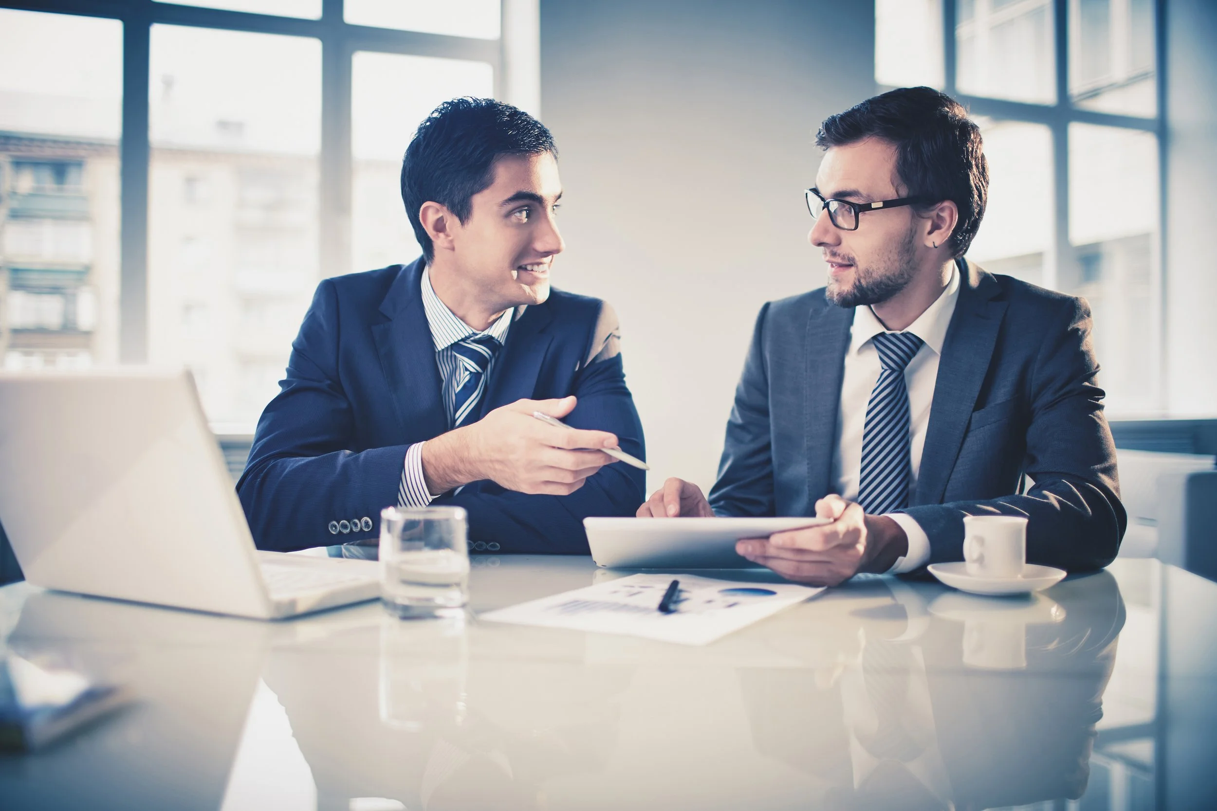 Two men in business suits talking and smiling during a meeting in a bright office with large windows.