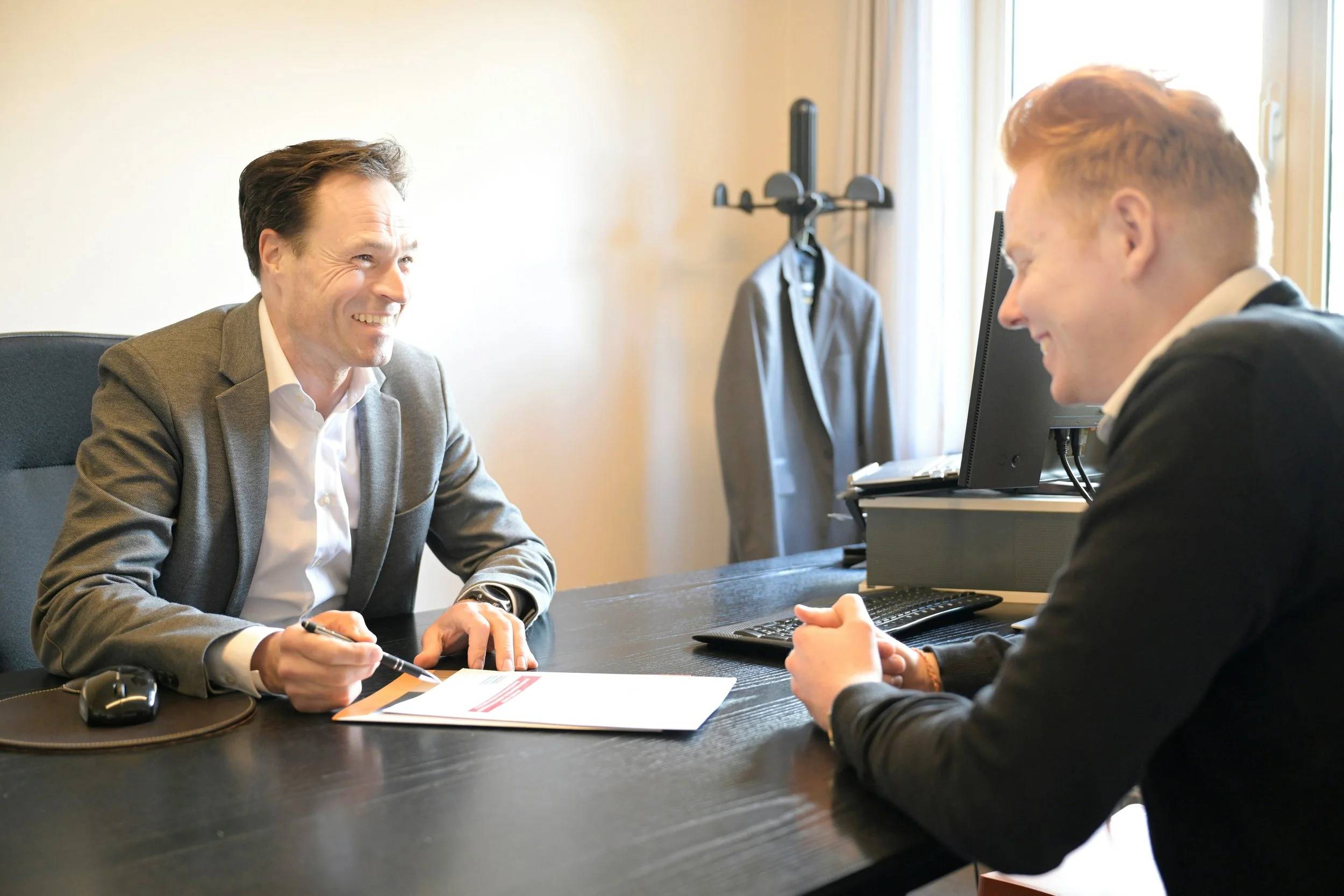 Two men in business attire engaging in a conversation across a desk in an office, with one holding a pen and the other smiling.
