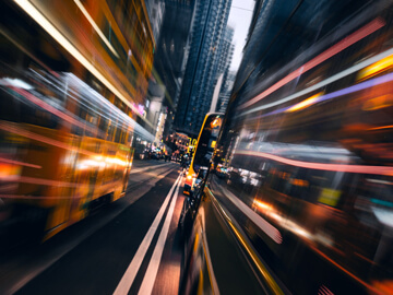 Blurred city street at night with yellow buses and tall buildings.