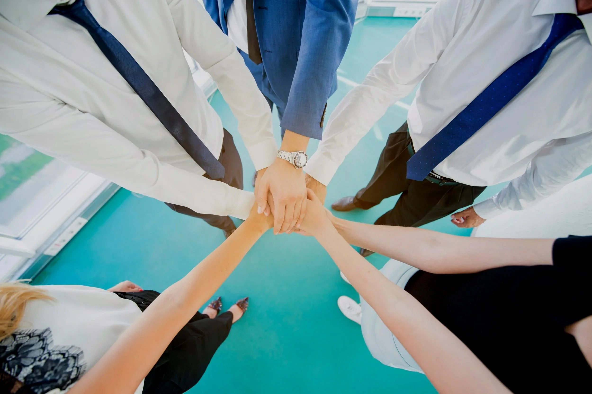Group of professionals in business attire stacking their hands together in a team-building gesture, seen from above.
