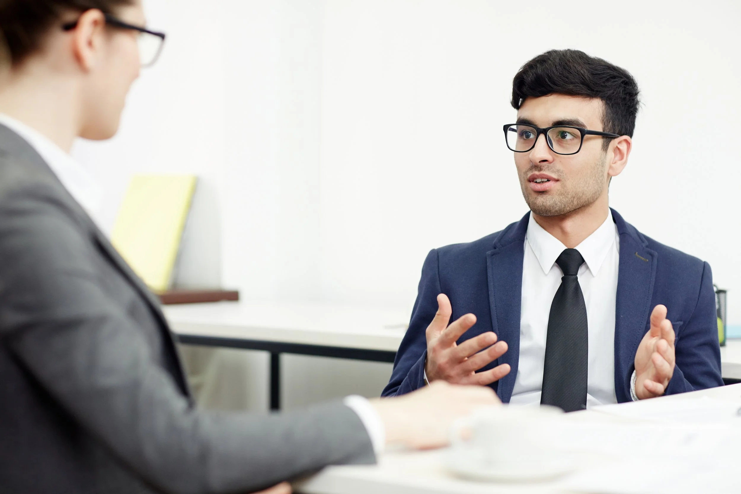 Two men in business attire and glasses are engaged in conversation, sitting at a table in a modern office with a plain white wall background.