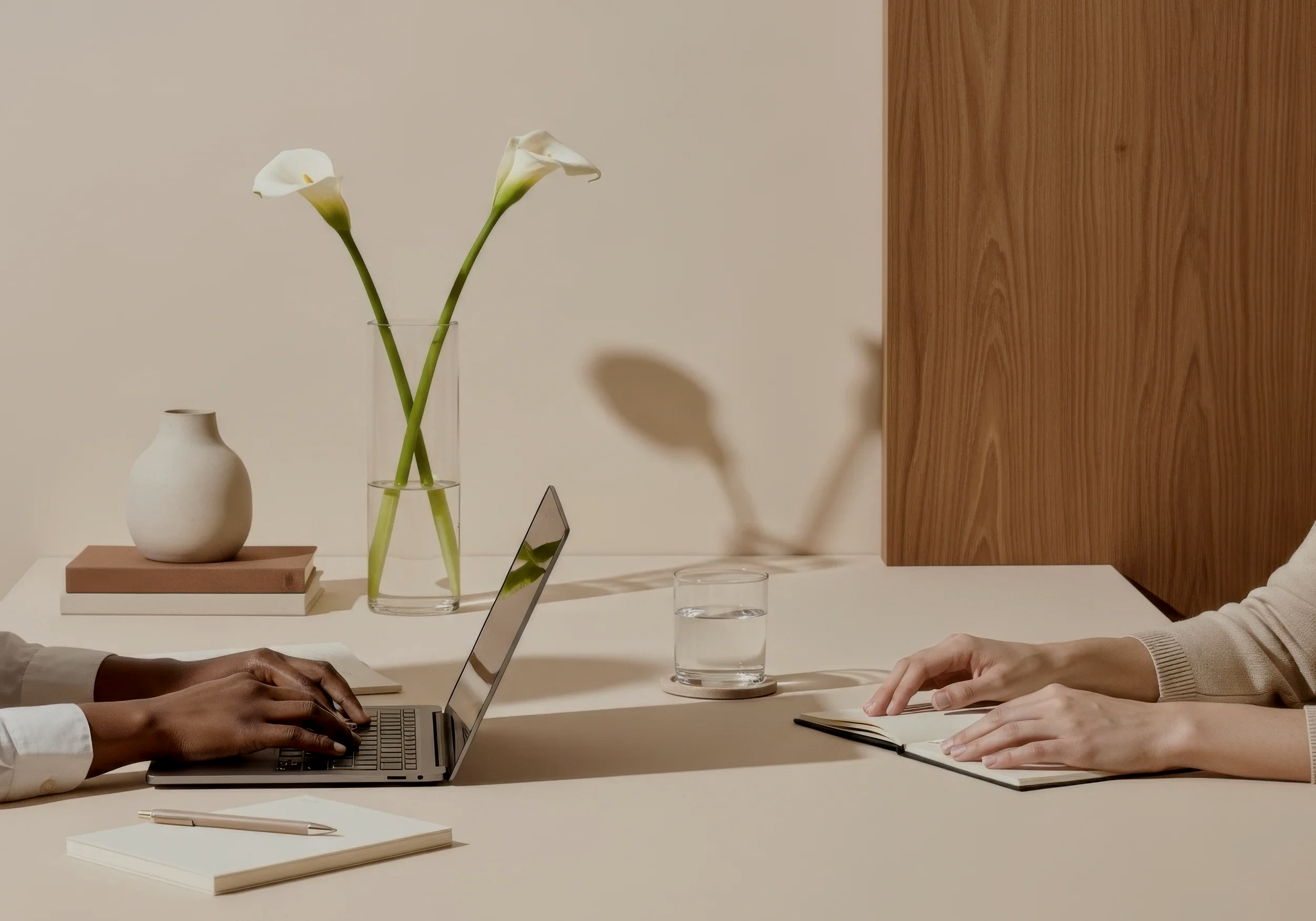 Two people sitting at a table with laptops and notebooks, having a meeting. There are vases with calla lilies, a small white vase, and a closed laptop on the table.