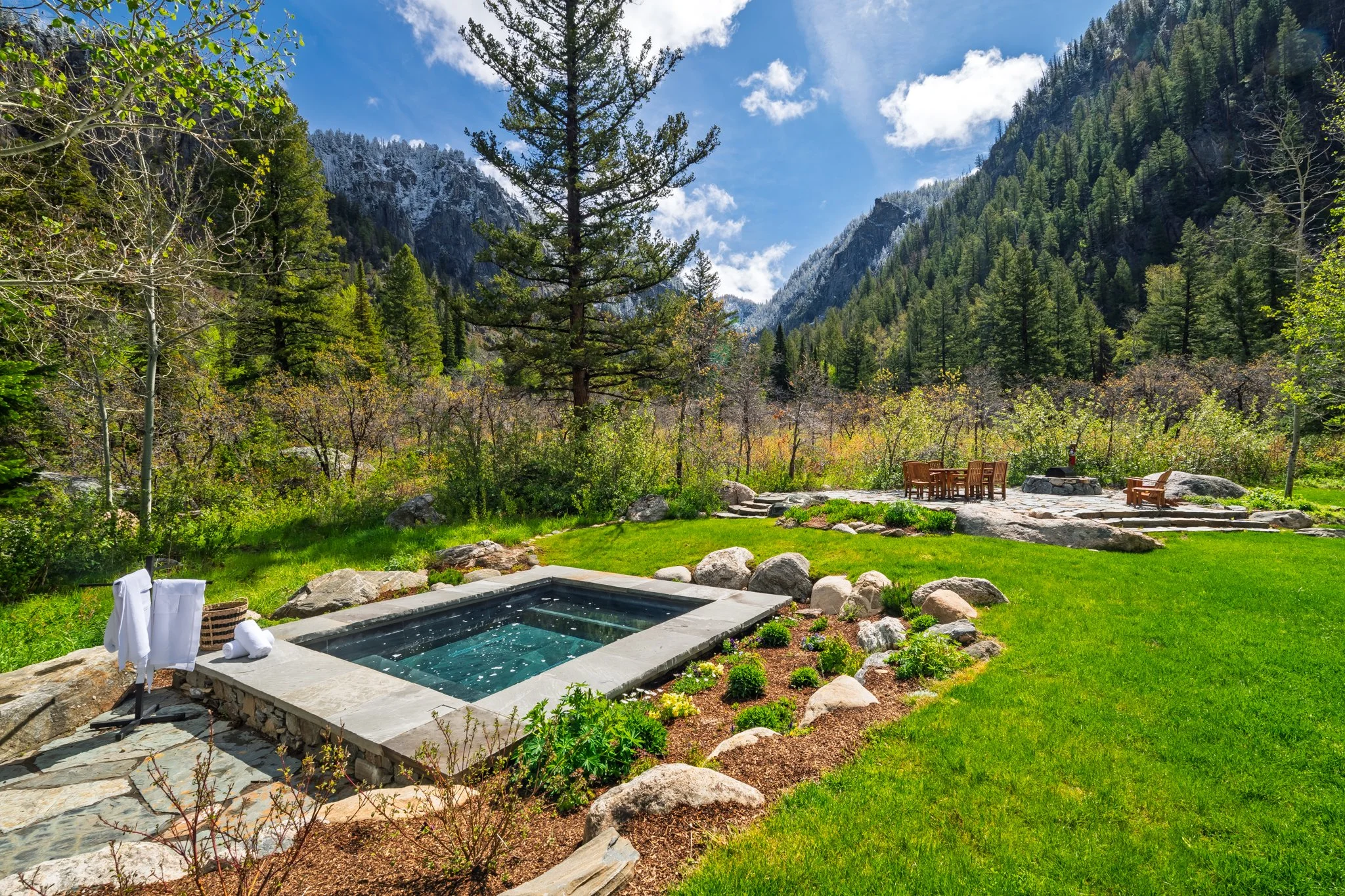 Backyard with a small pool, patio furniture, surrounded by green grass, rocks, trees, and mountains under a partly cloudy sky.