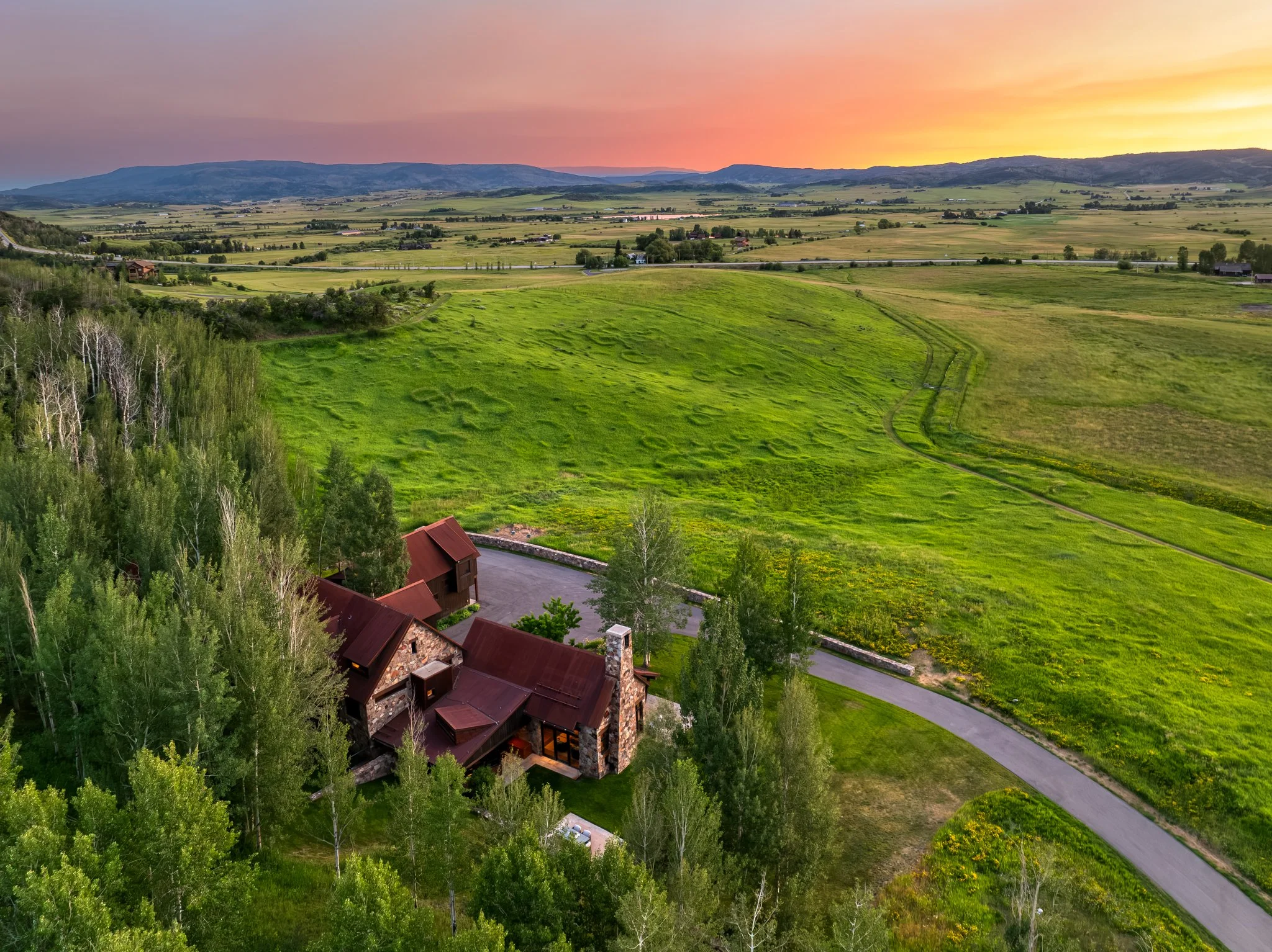 A rural landscape at sunset featuring rolling green fields, scattered houses, and distant mountains.